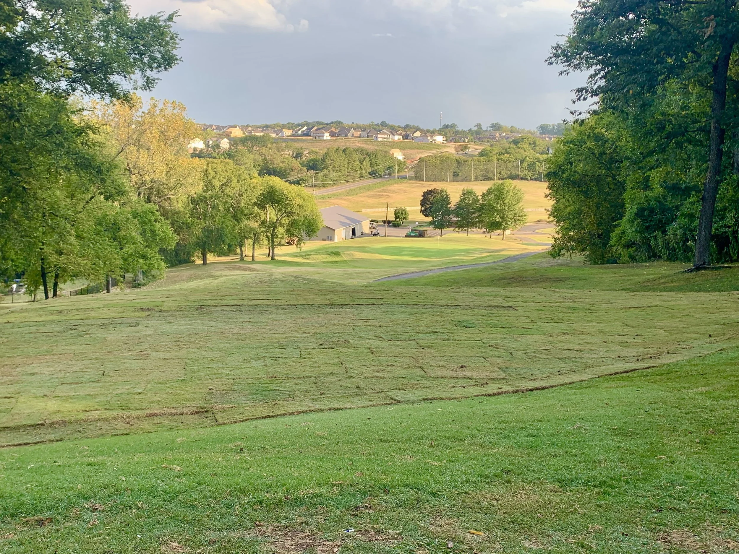 A golf course with well-maintained grass, surrounded by trees, and residential houses on a hill in the background under a partly cloudy sky.