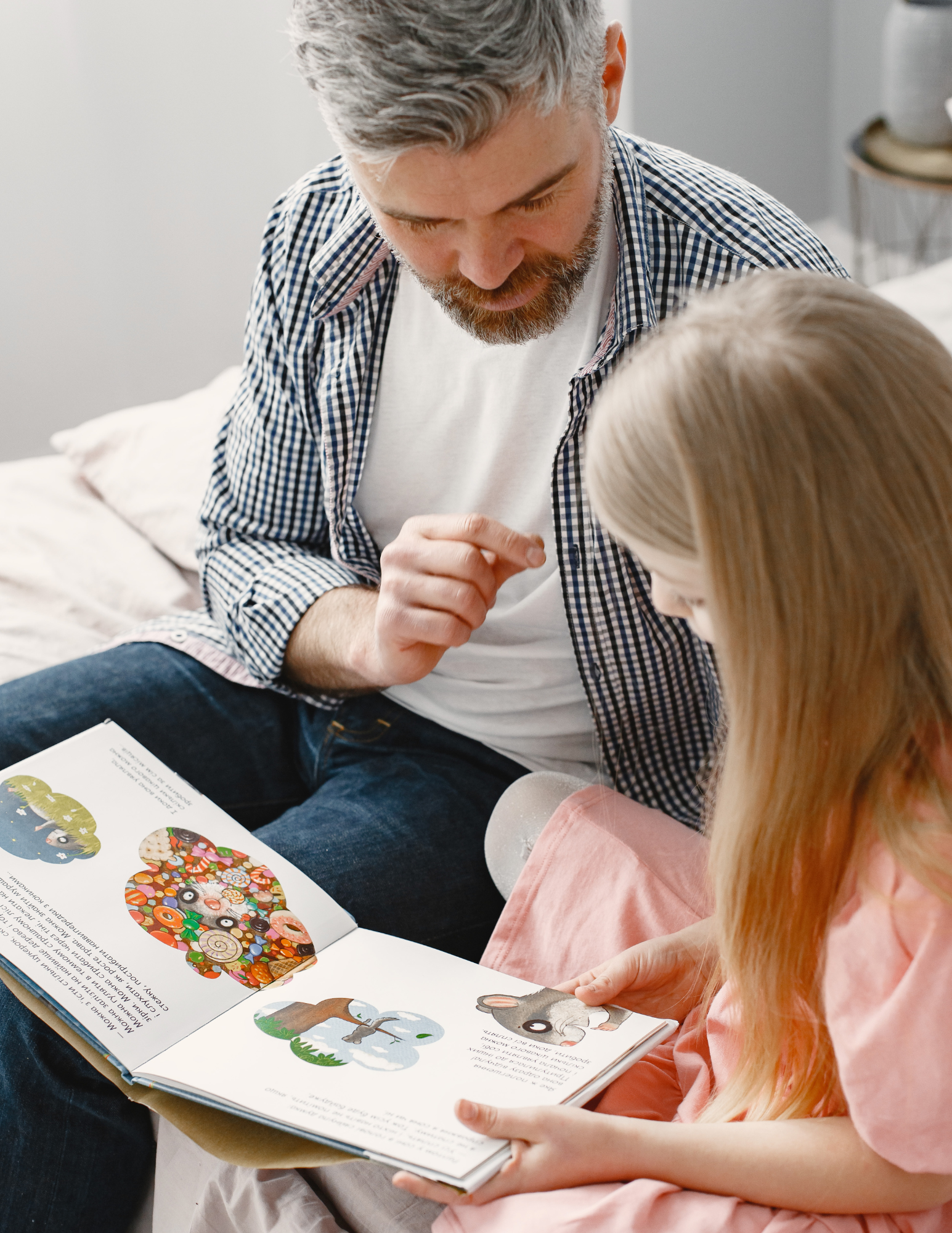 A man with gray hair and a beard is looking at a young girl with long blonde hair, who is reading a picture book together. The book features colorful illustrations of animals and objects.