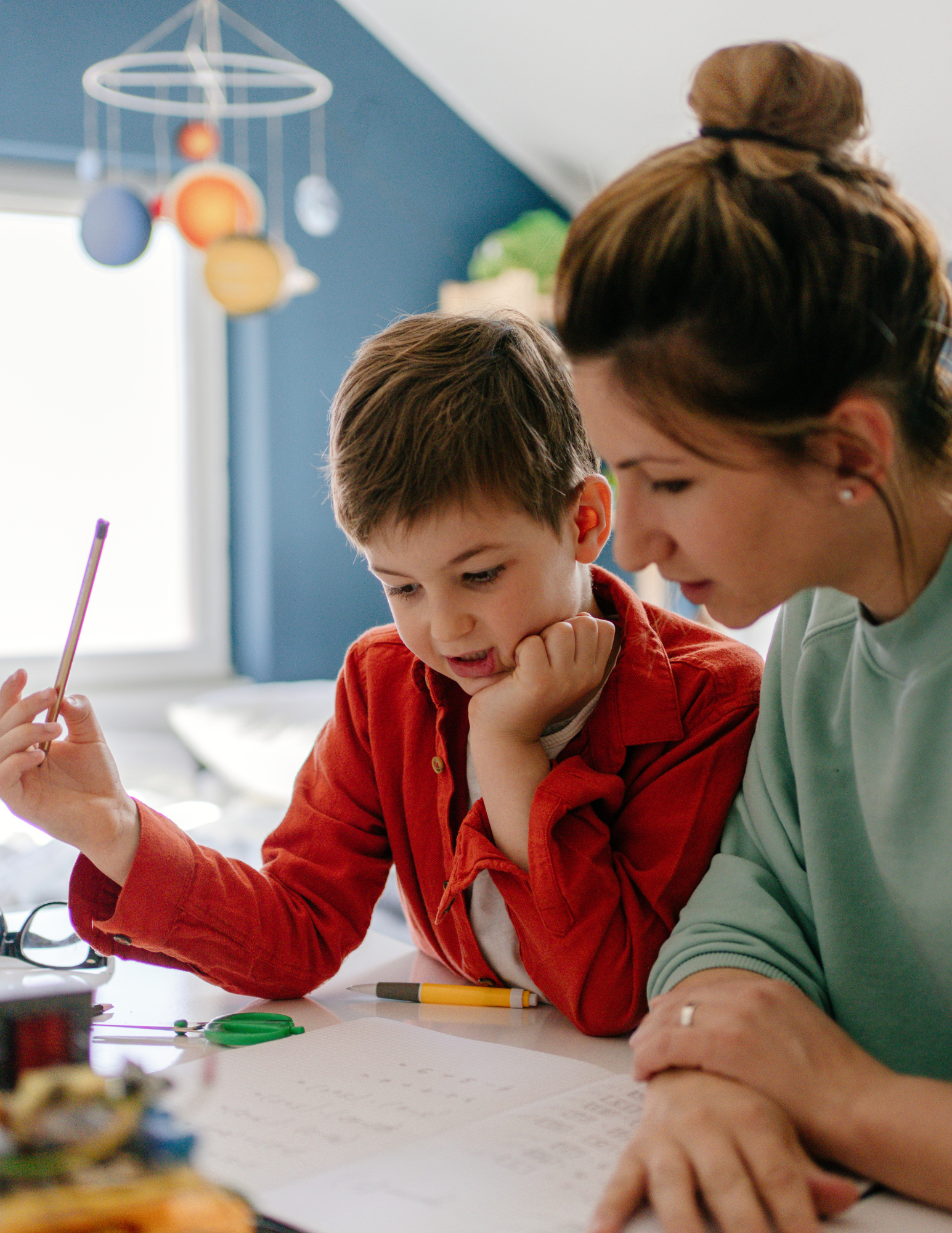 A woman and a young boy are sitting at a table, looking intently at a paper with writing. The woman has brown hair styled in a bun with a black scrunchie, and the boy has short brown hair. There are various school supplies, including a yellow pencil, visible on the table. In the background, a colorful hanging mobile and a window are visible.