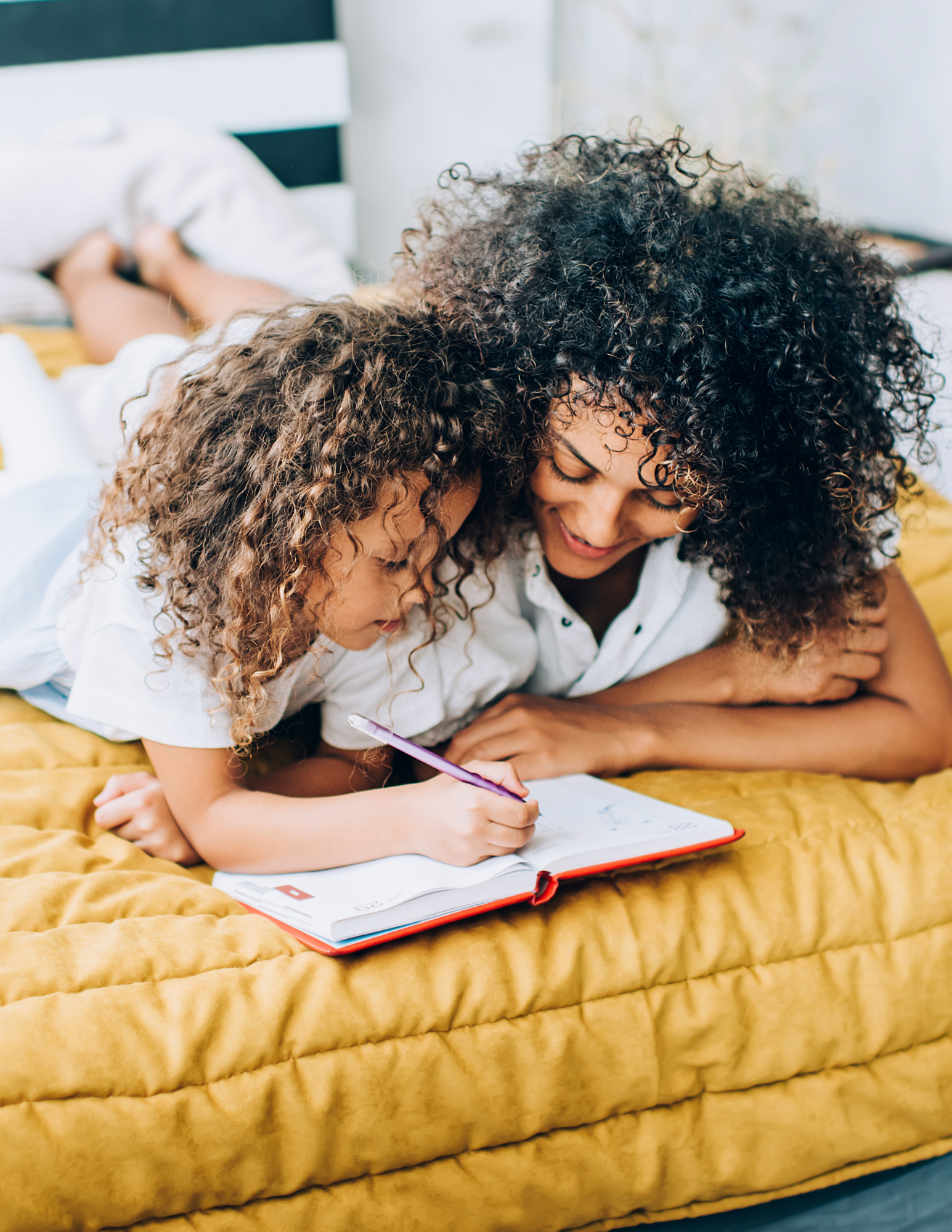 A woman and a young girl, both with curly hair, are lying on a yellow quilted bedspread, looking at and writing in a red notebook together.