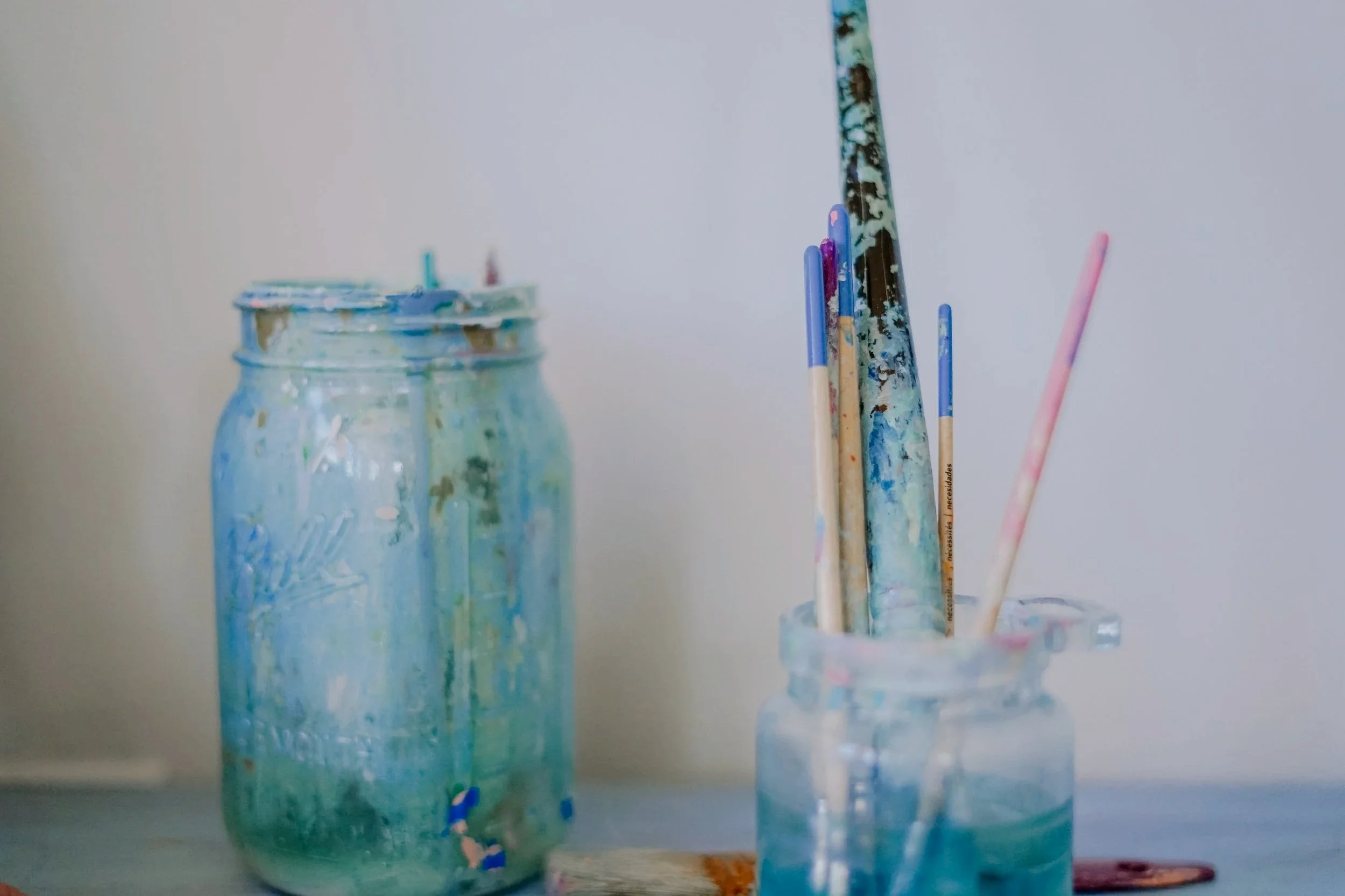A jar filled with water and painted with various colors, and a glass container holding several painted and stained wooden sticks or brushes with colorful tips, possibly used for painting or artistic purposes.