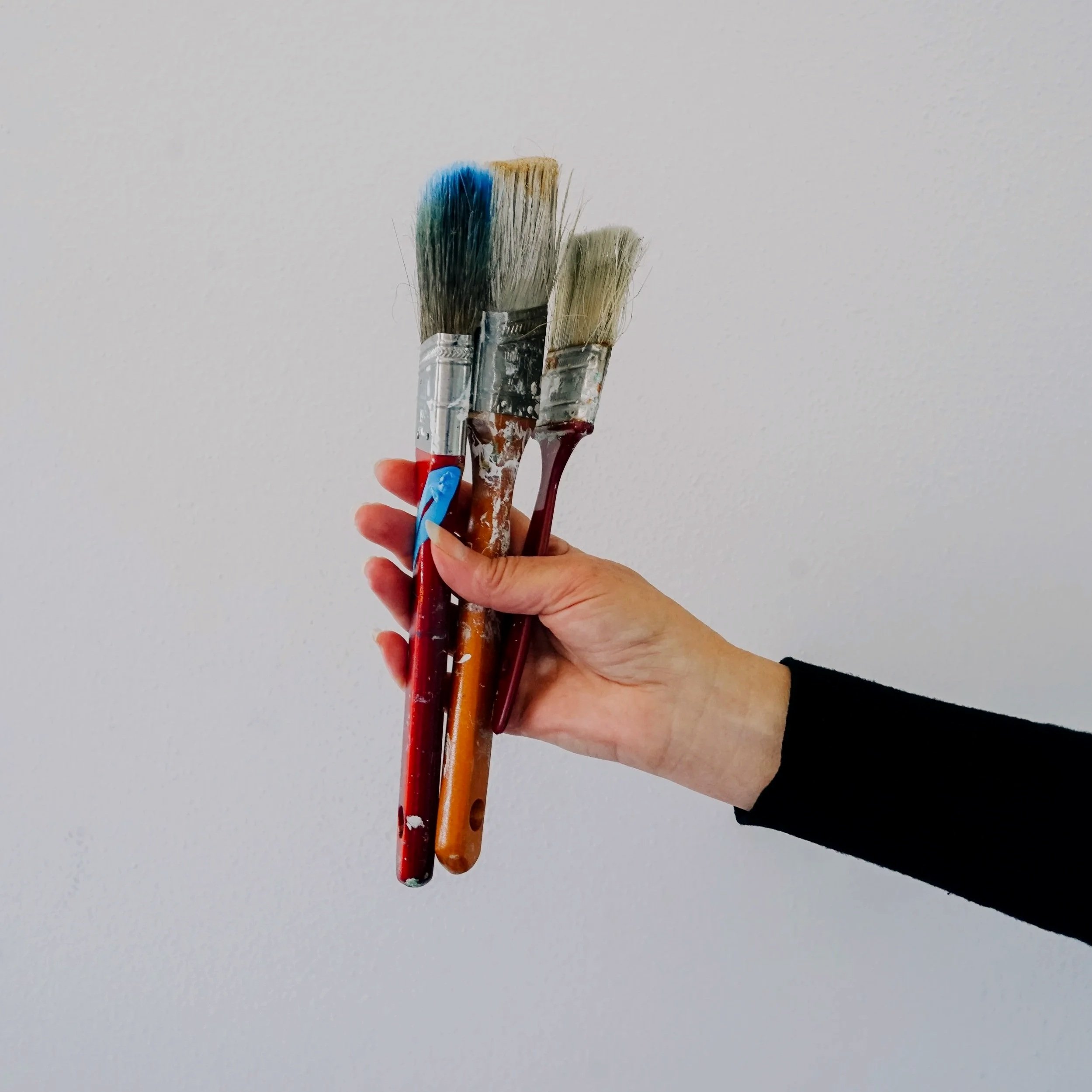 A hand holding three paintbrushes with paint on the handles against a plain white background.