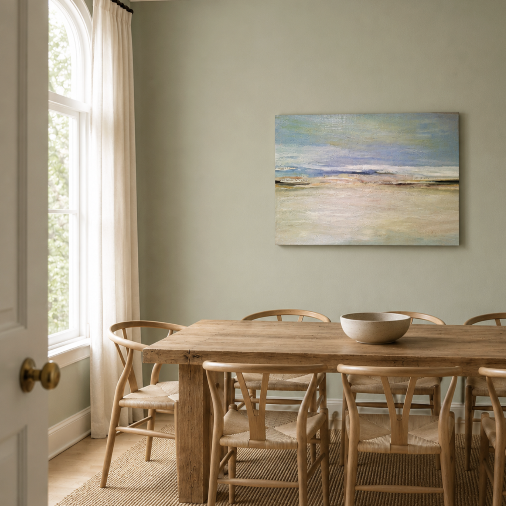 Dining room with wooden table, chairs, a bowl, large window with curtains, and a landscape painting on the wall.