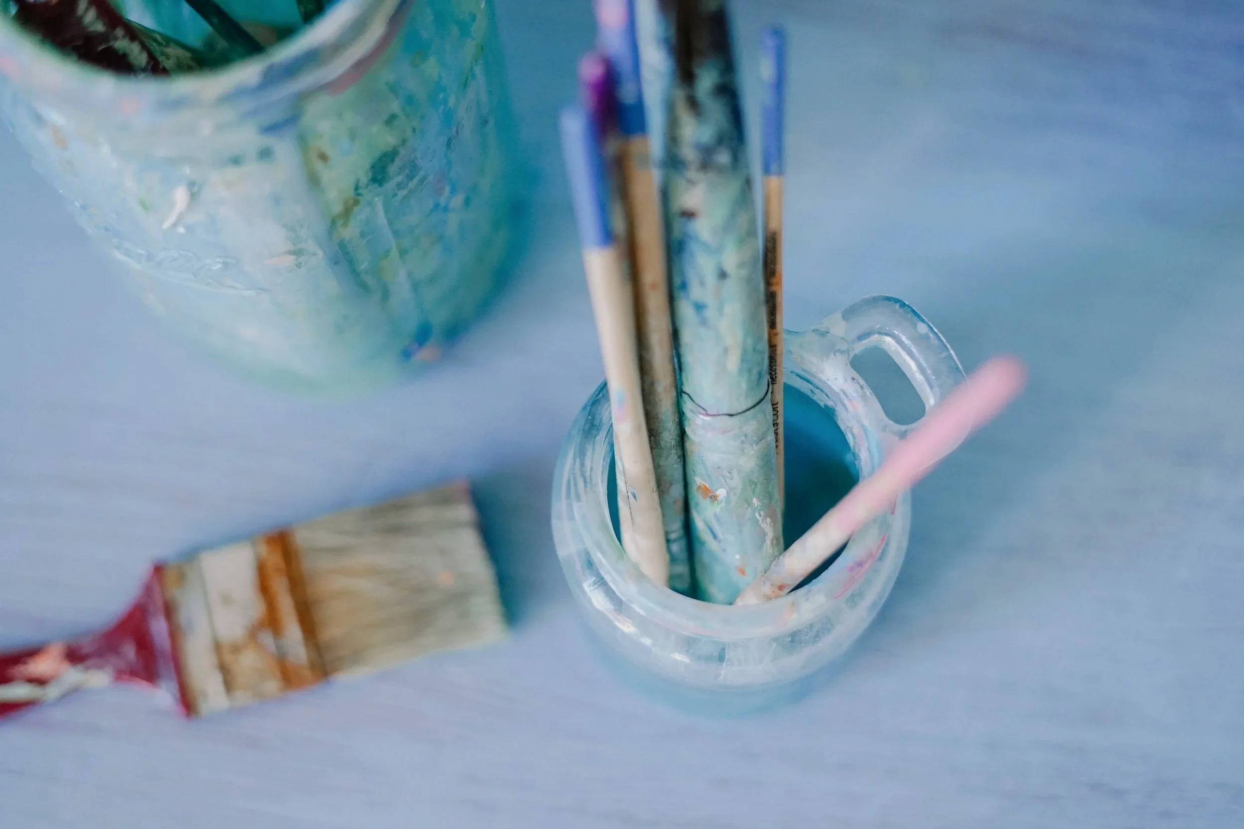 A clear glass jar holding paintbrushes on a blue surface with a blurred colorful background.