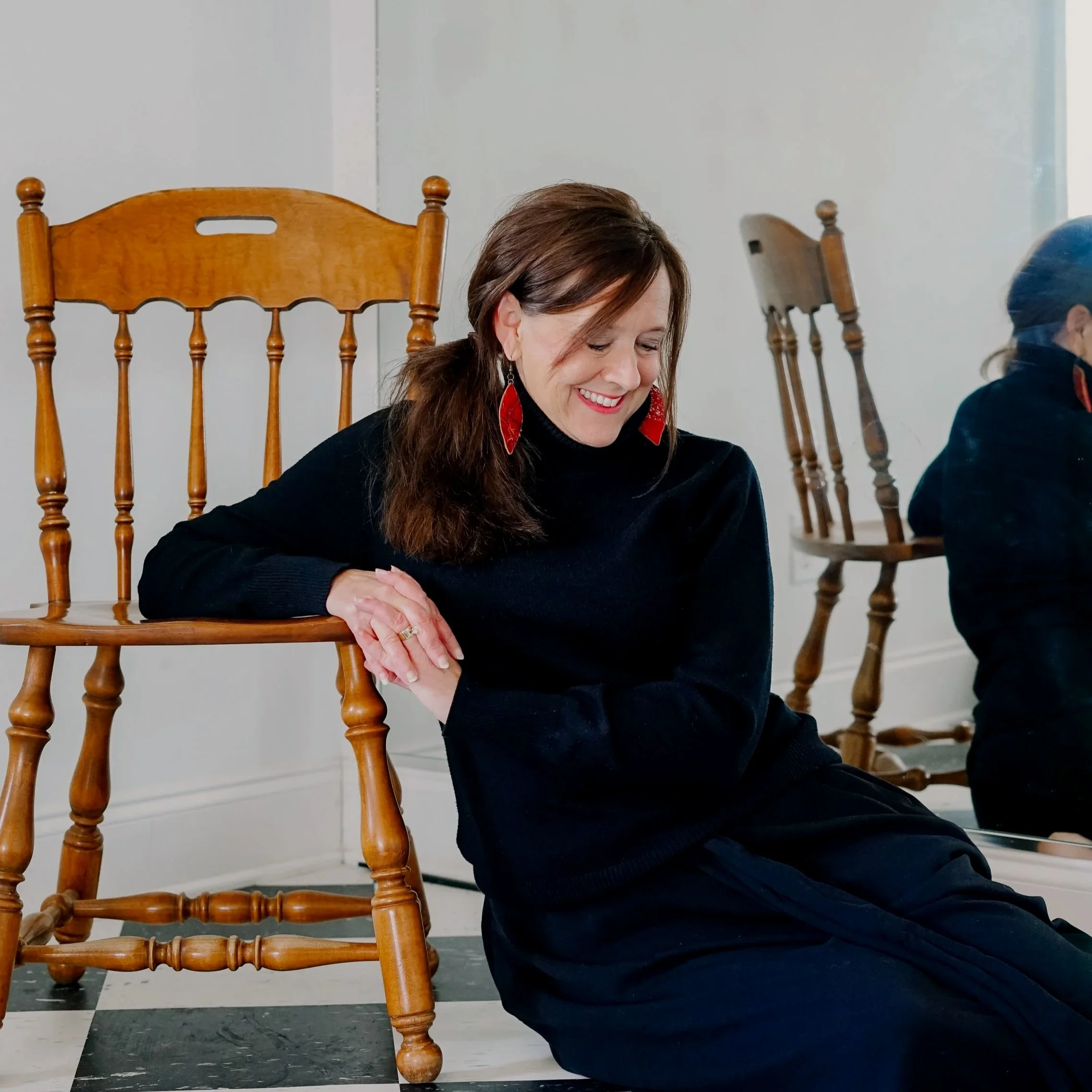 A woman kneeling on the floor with her head bowed and eyes closed, resting her arms on a wooden chair with a high back and turned spindles. The background includes additional wooden chairs and a white wall.