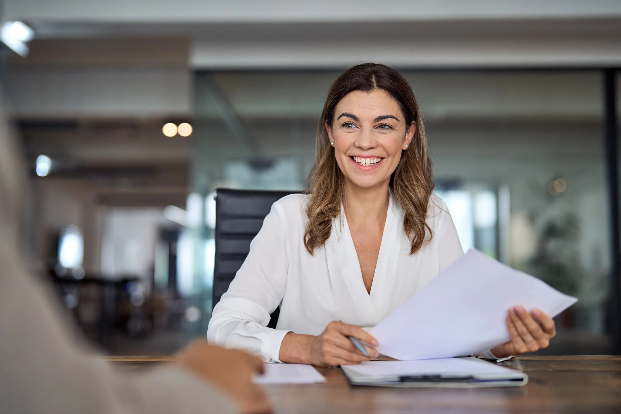 A smiling woman with brown hair in a white blazer sitting at a desk with papers, in a modern office environment.