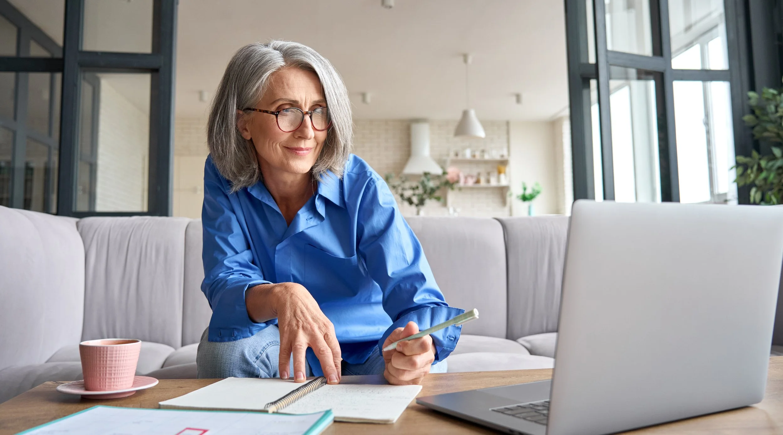 An older woman with gray hair, glasses, wearing a blue shirt, sitting on a beige couch in a bright, modern room. She is using a smartphone, with papers, a notebook, a cup, and a laptop on the table in front of her.