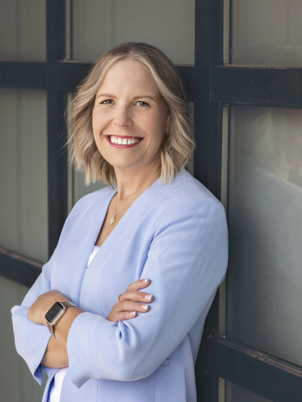 Smiling woman with shoulder-length blonde hair and blue eyes, wearing a light blue blazer, a necklace, a watch, and a ring, standing with arms crossed against a metal and glass window wall.