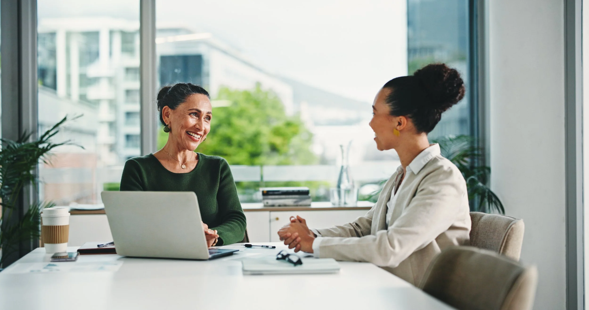 Two women having a conversation in a bright, modern office with large windows, a laptop, and a coffee cup on the table.