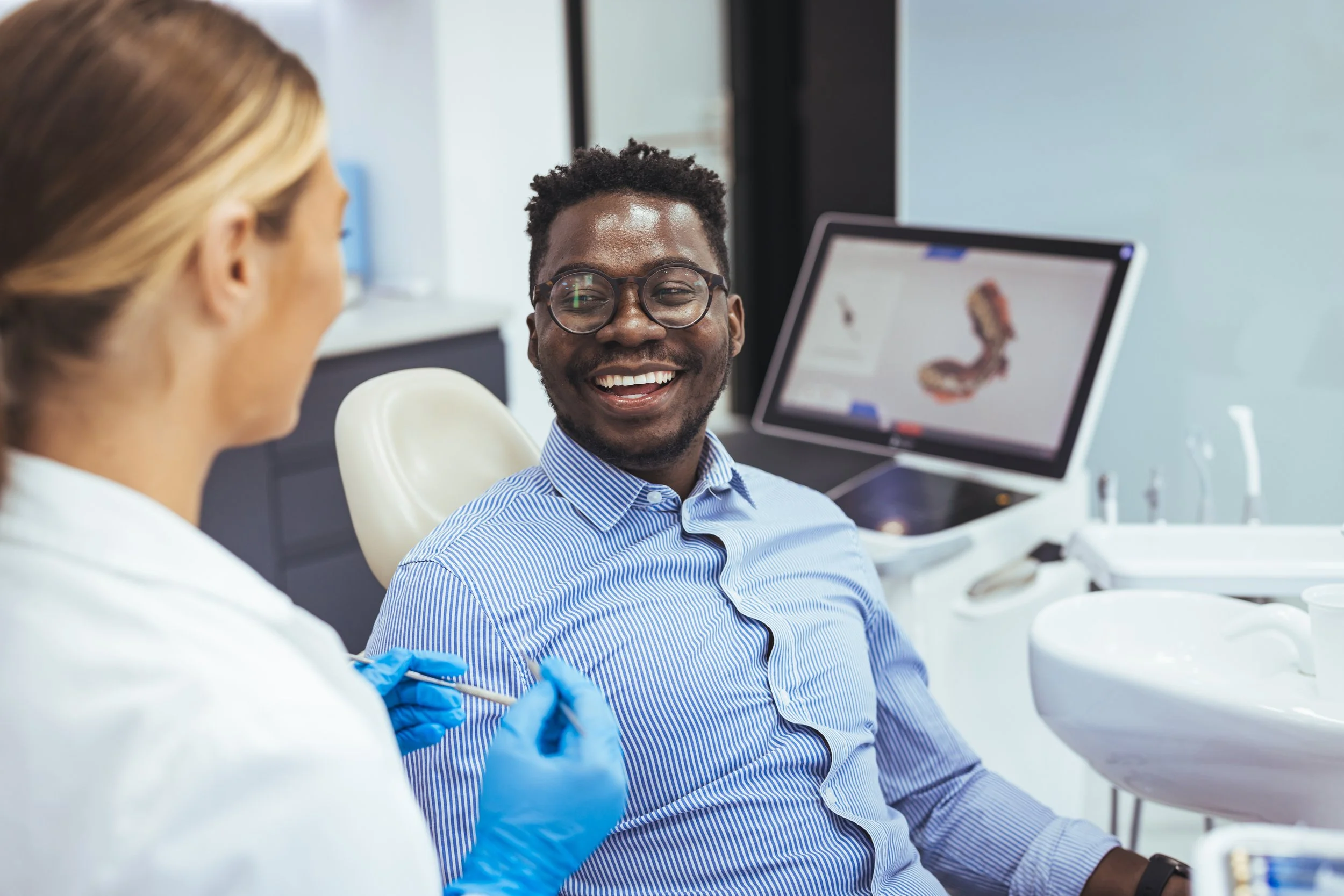 A man with glasses smiling while sitting in a dental office, talking to a dental professional seated nearby who is wearing blue gloves.