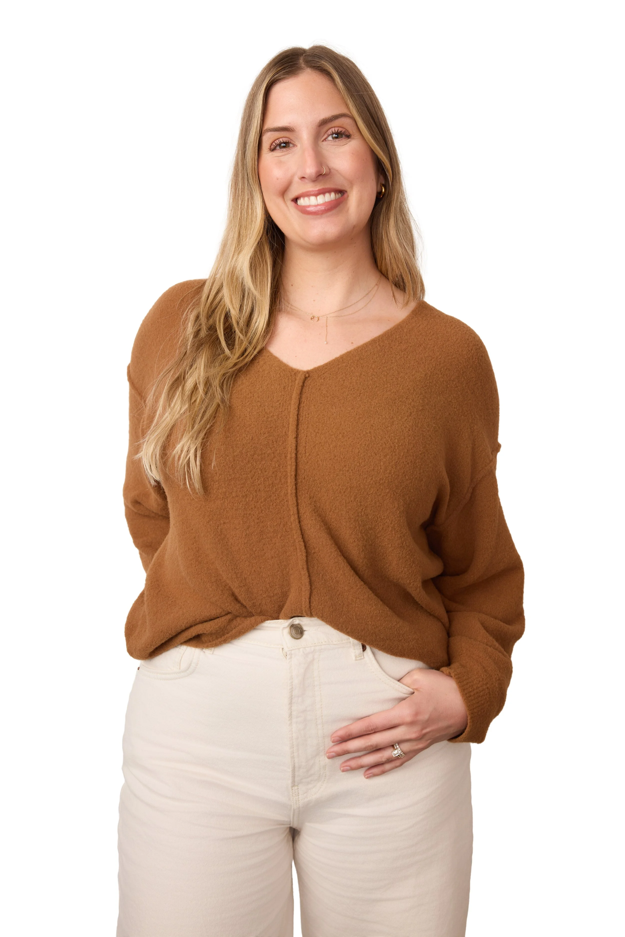 A woman with long wavy blonde hair, wearing a brown sweater and cream pants, smiling and standing with her right hand on her hip against a white background.