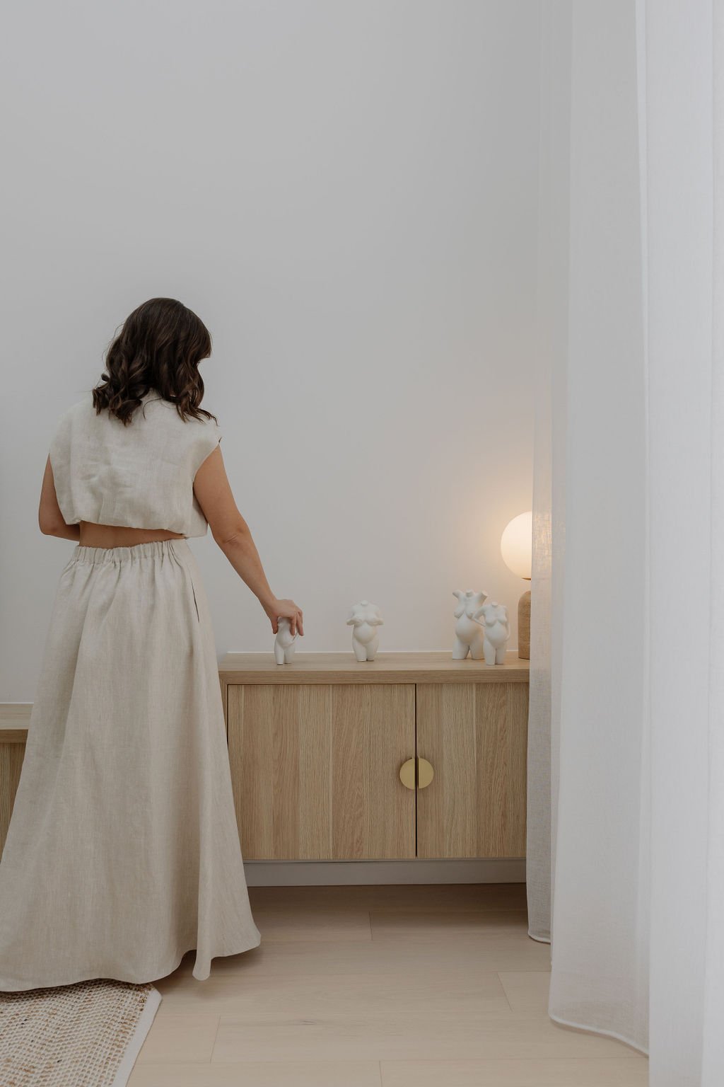 A woman in beige clothing placing a white pregnancy sculpture or bust or statue, on a minimalistic light wood console table in a room with white walls and a small round light fixture.