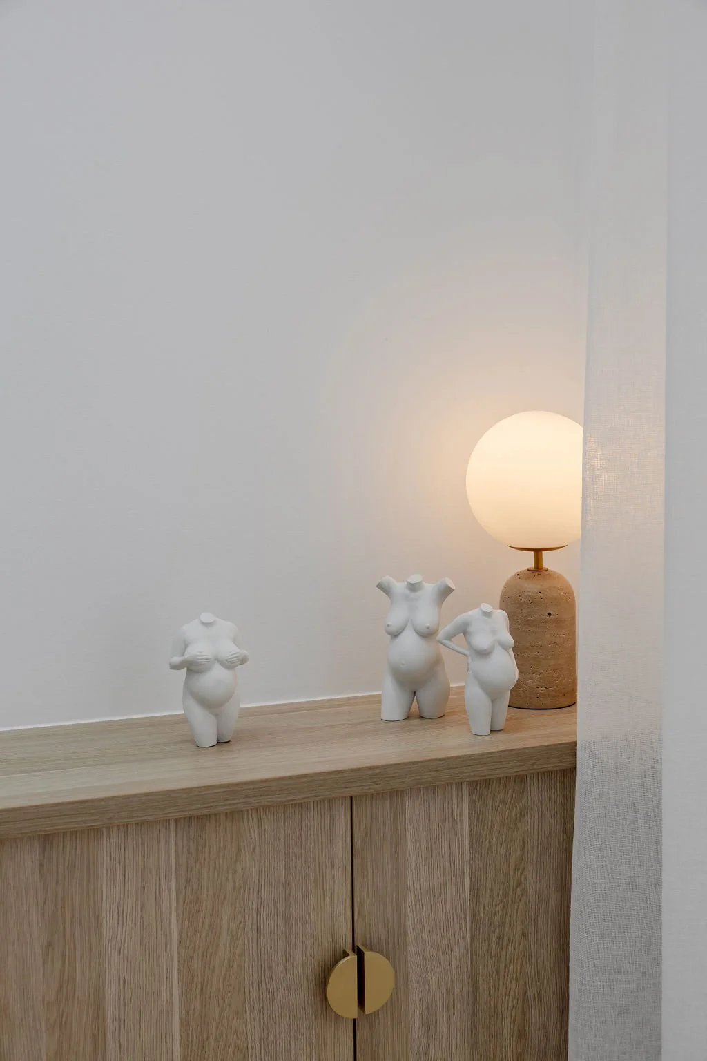 Decorative white sculptures of female torsos on a wooden surface with a lit table lamp next to them.