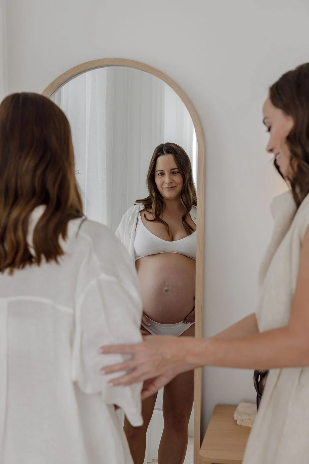 A pregnant woman looking at herself in a mirror, smiling gently, with her reflection visible.