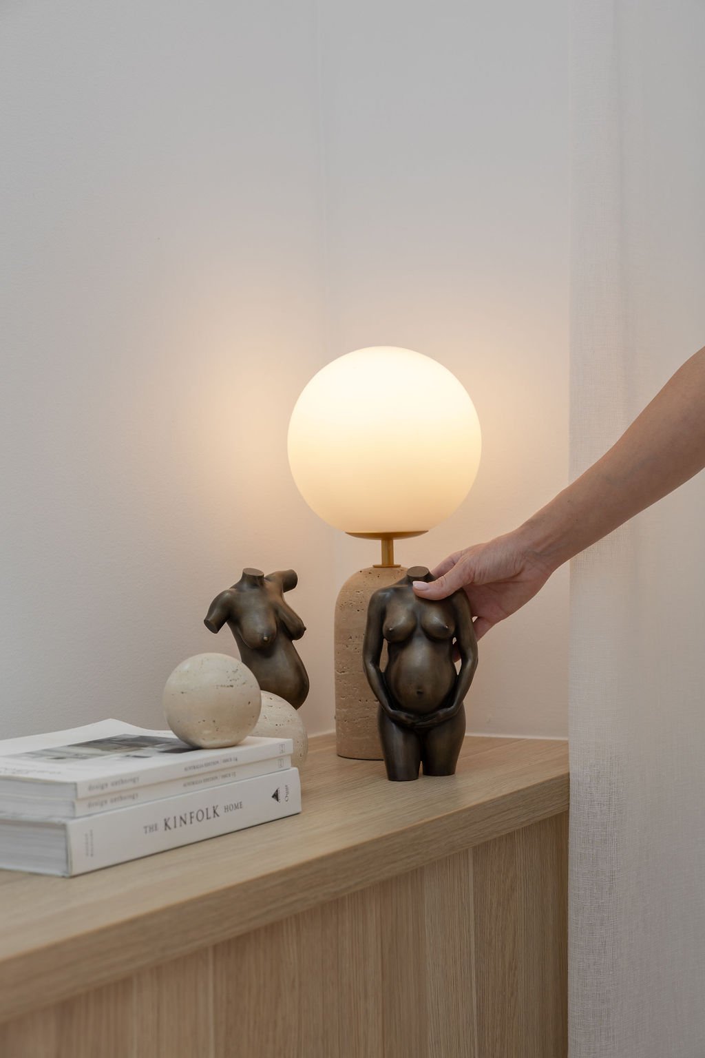 A wooden table with decorative sculptures of female torsos, a spherical object, and reading materials, with a lamp featuring a spherical light shade, against a plain wall.