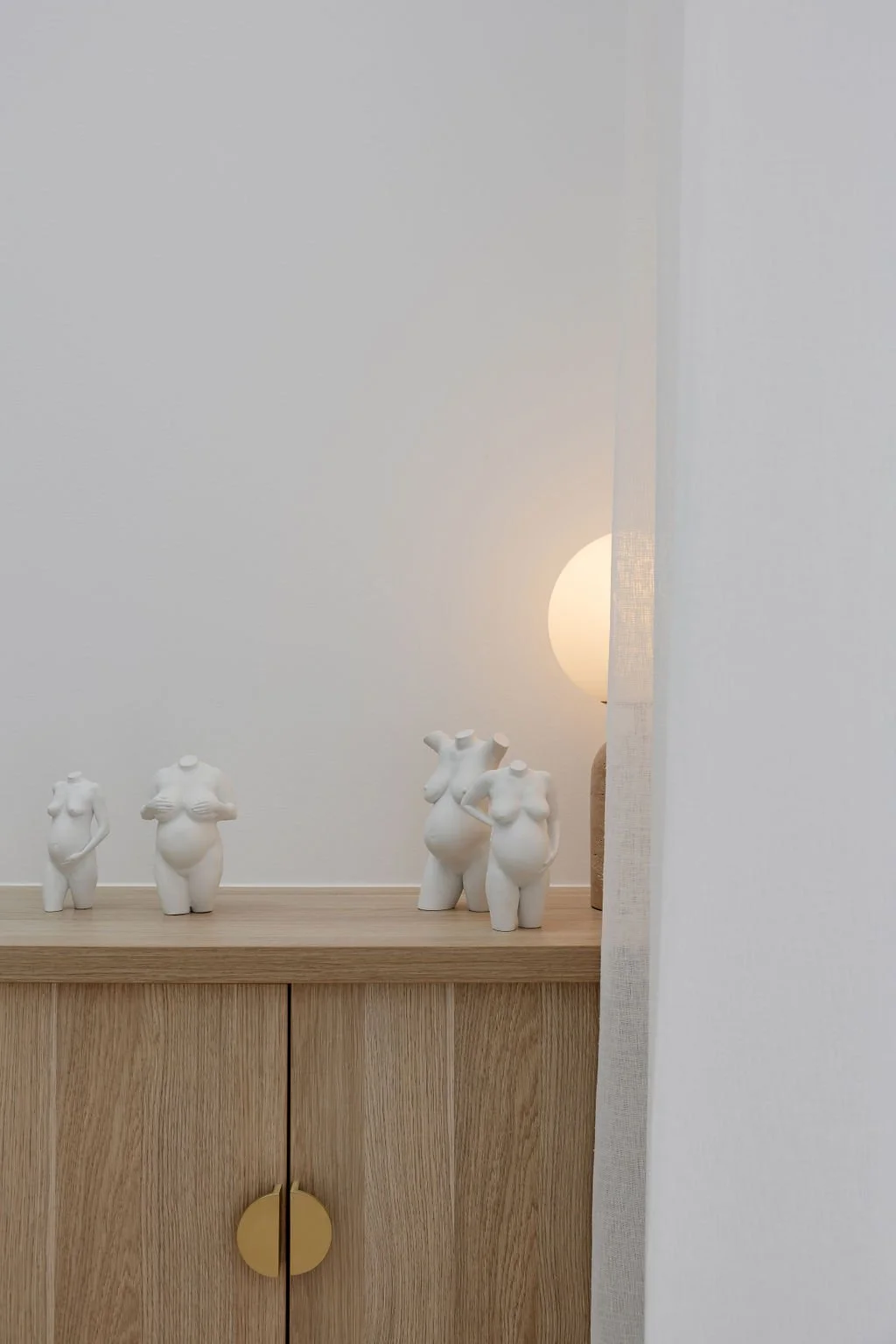 Decorative white sculptures of female torsos and bodies on a wooden surface, with a luminescent light in the background and partially visible curtain.