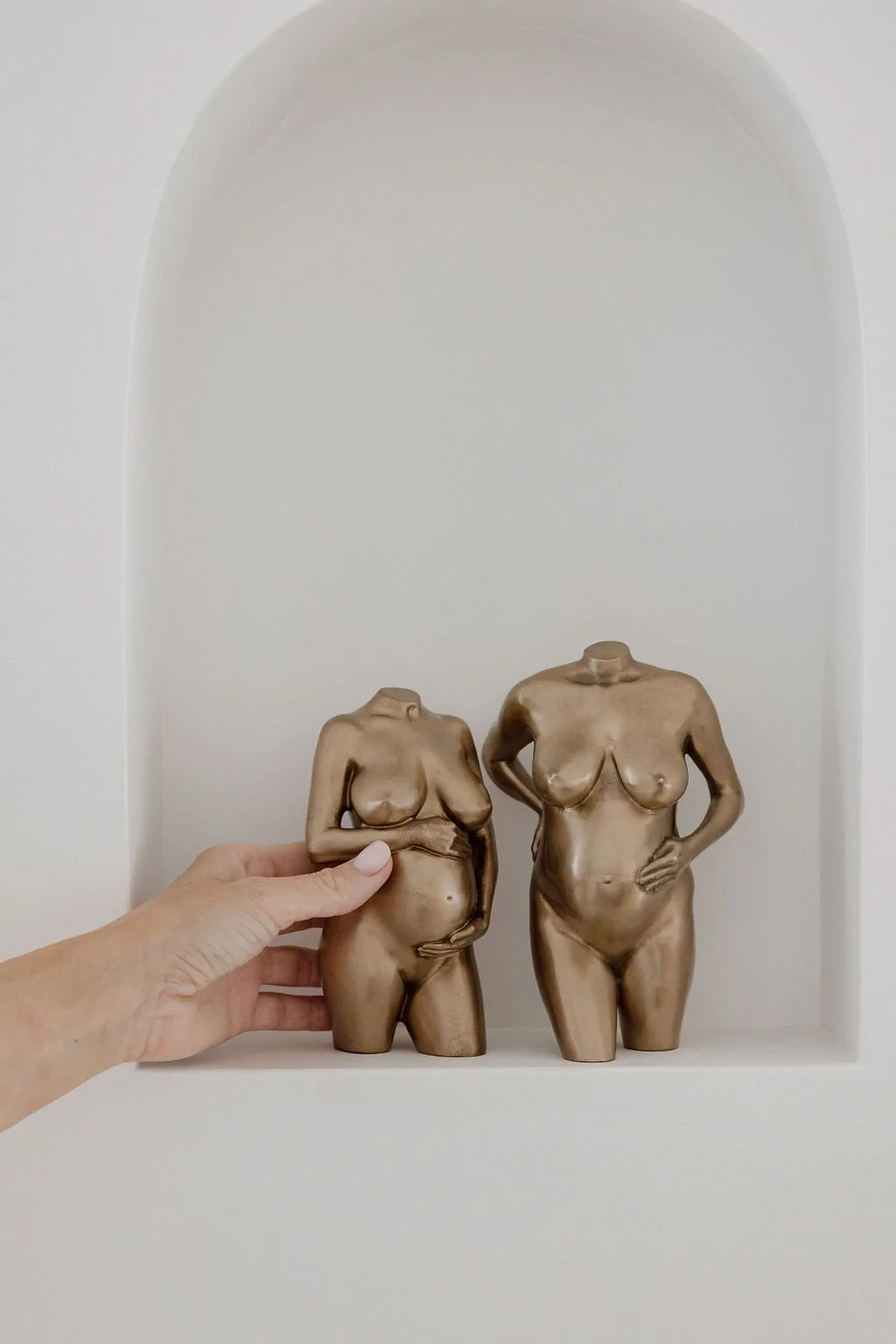 Two bronze sculptures of female torsos, one smaller and one larger, placed on a shelf against a white background, with a hand holding the smaller sculpture.