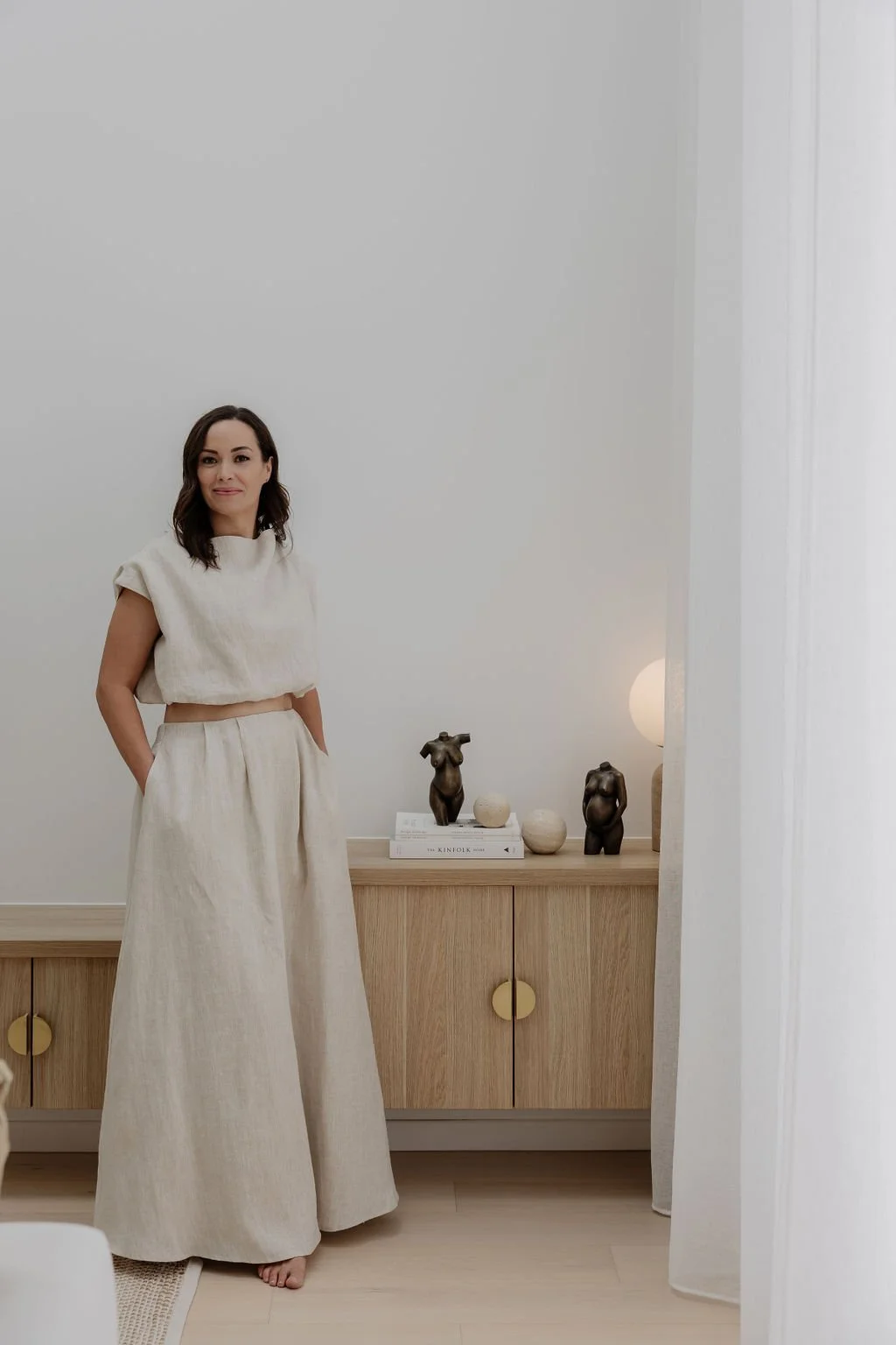 Woman standing in a minimalist, light-colored room with a wooden console table displaying small sculptures and books.