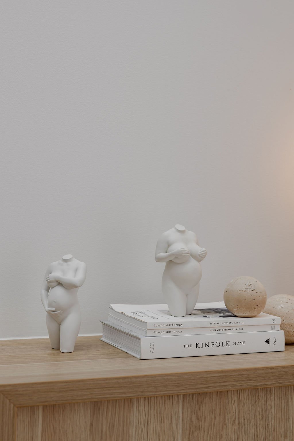 Two sculptures with arms crossed over their chests, placed on a wooden surface alongside a stack of books titled 'Design Anthology' and 'The Kinfolk Home', with two round textured stones on the right.