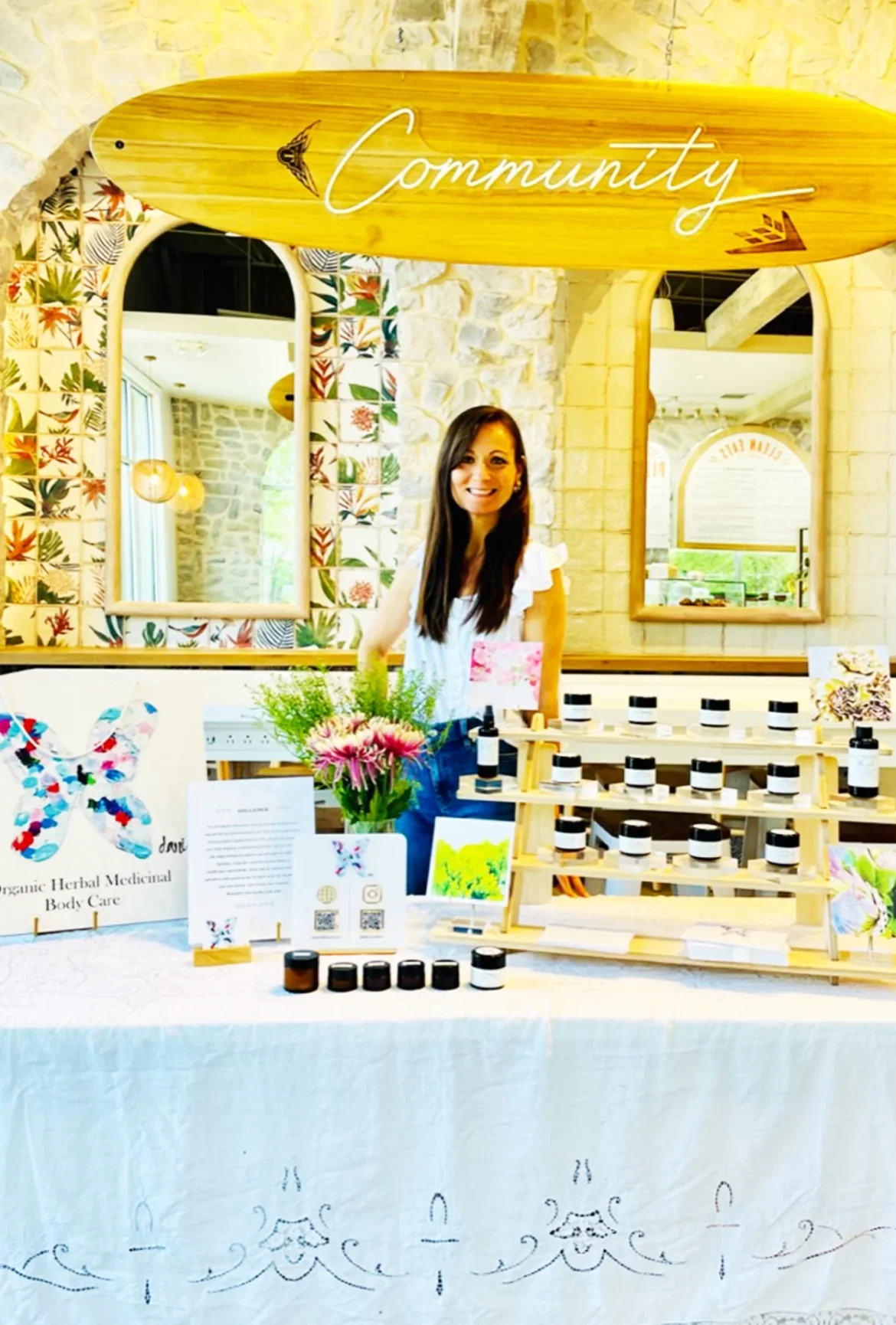 Woman smiling behind a table at a booth with homemade herbal products, flowers, and decorative butterfly art, with a sign that reads 'Community' above.