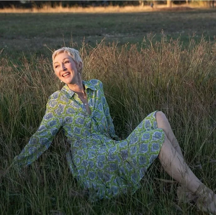 A woman sitting in tall grass in a field, smiling and looking to the side, wearing a patterned dress.