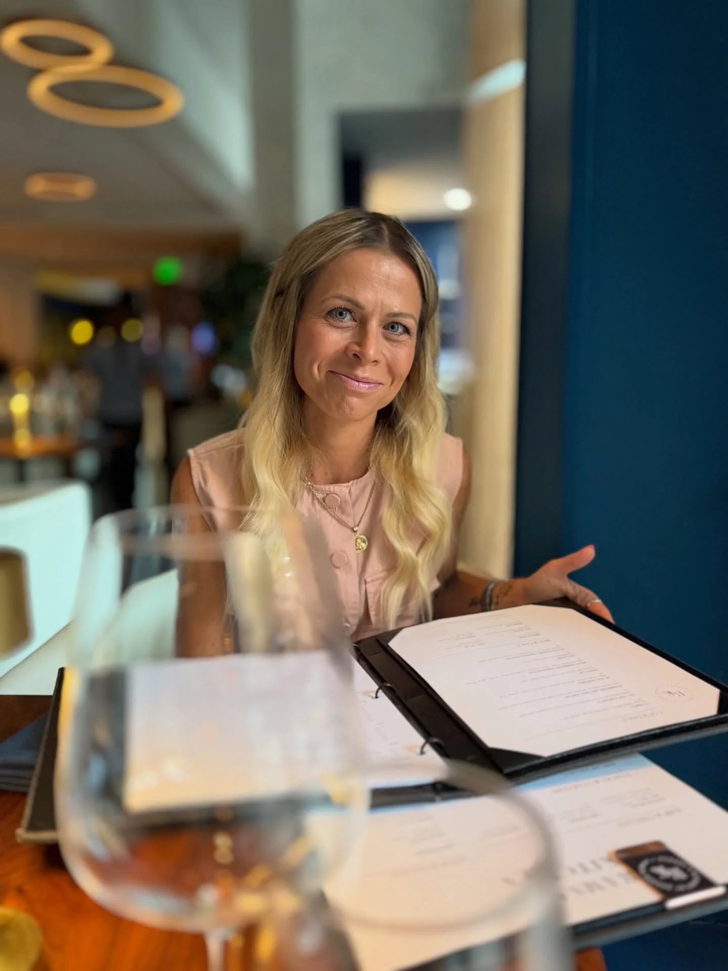 A woman with blonde hair sitting at a restaurant table, smiling, with a menu in front of her. There is a glass of water and some drinks on the table.