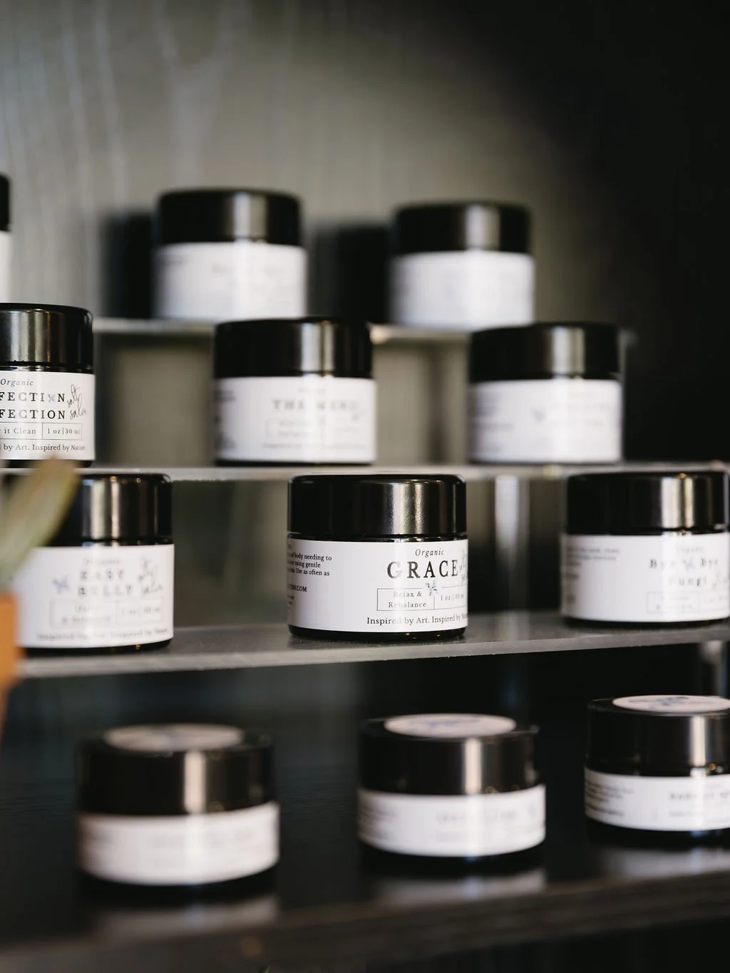 Shelf with small black jars labeled with white labels, some jars are in foreground and some in background, on a metal shelf, with a gray wall behind.