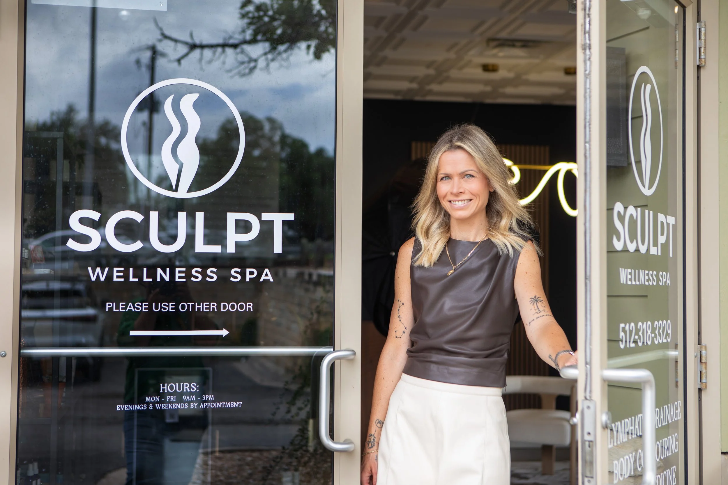 A woman with wavy blonde hair, wearing a black sleeveless top and a pearl necklace, smiles as she stands in front of the entrance to a wellness spa called 'SCULPT FITNESS & WELLNESS SPA'. The spa's glass door has the spa's logo and name, and a sign listing the hours of operation.