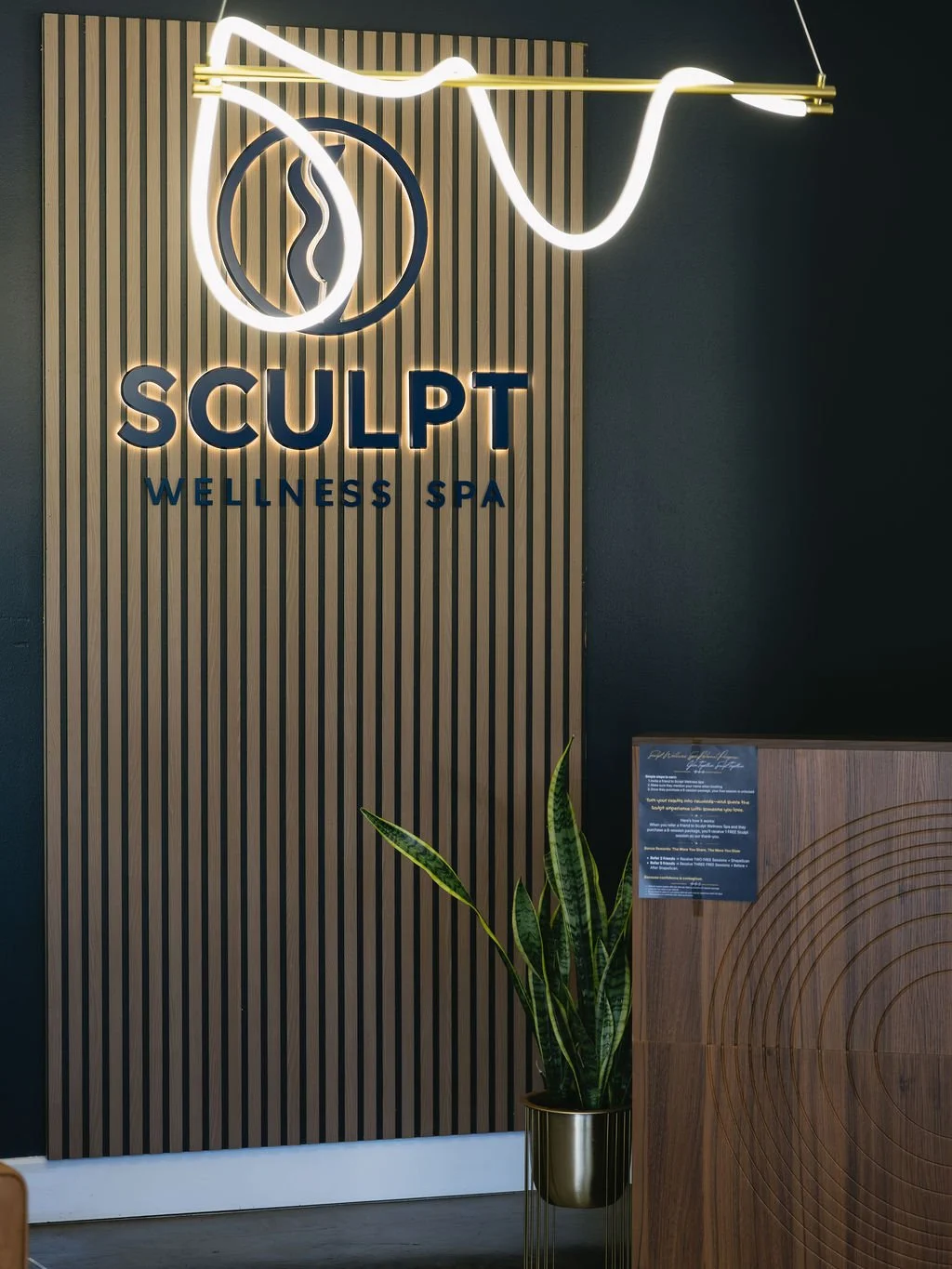 Neon sign reading "Sculpt Wellness Spa" on a wooden slat wall with a snake icon, a potted plant, and a wooden reception desk.