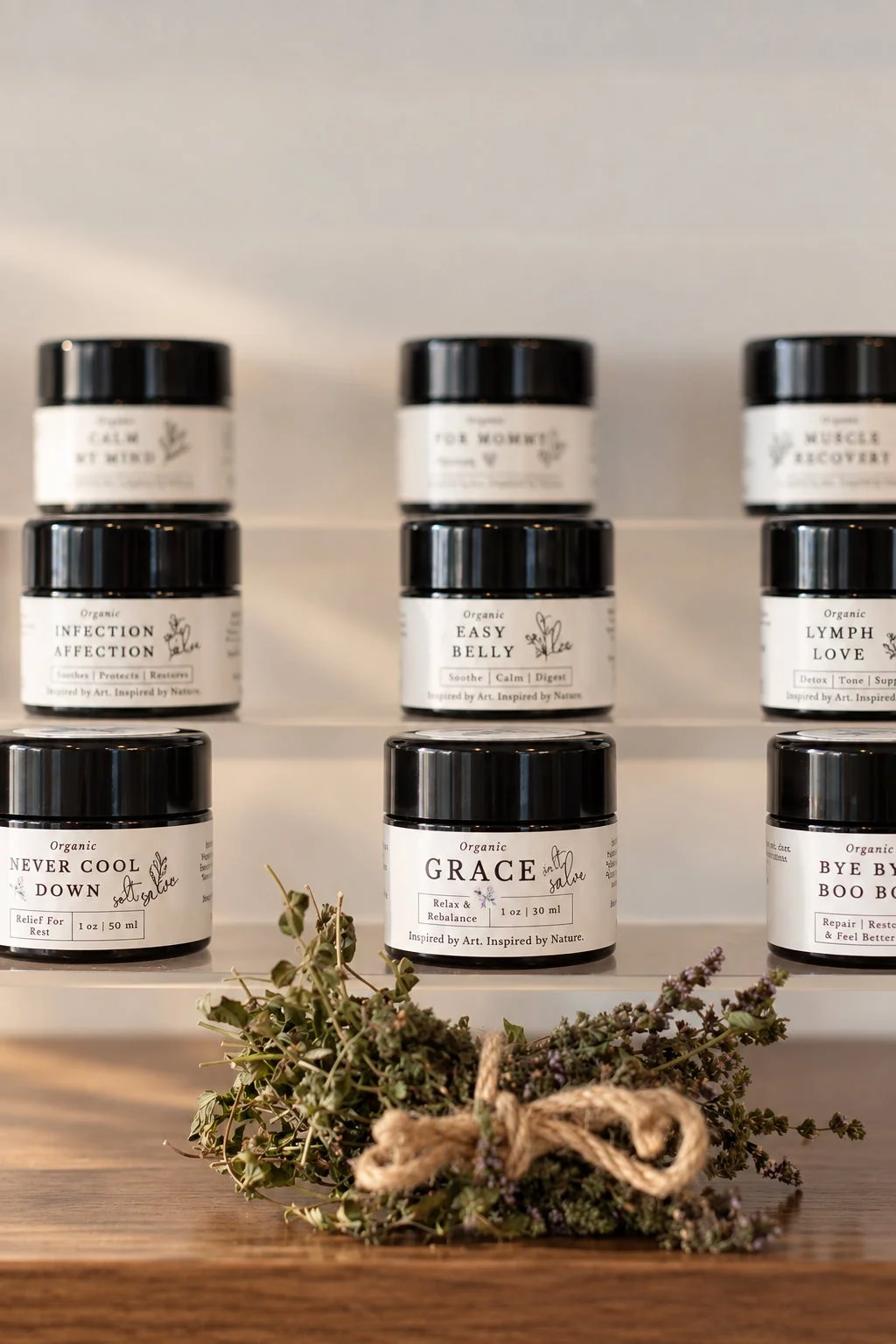 Assorted organic skincare or wellness products in black jars with white labels on a white shelf, with a small bouquet of dried herbs or flowers with a twine bow in front on a wooden surface.
