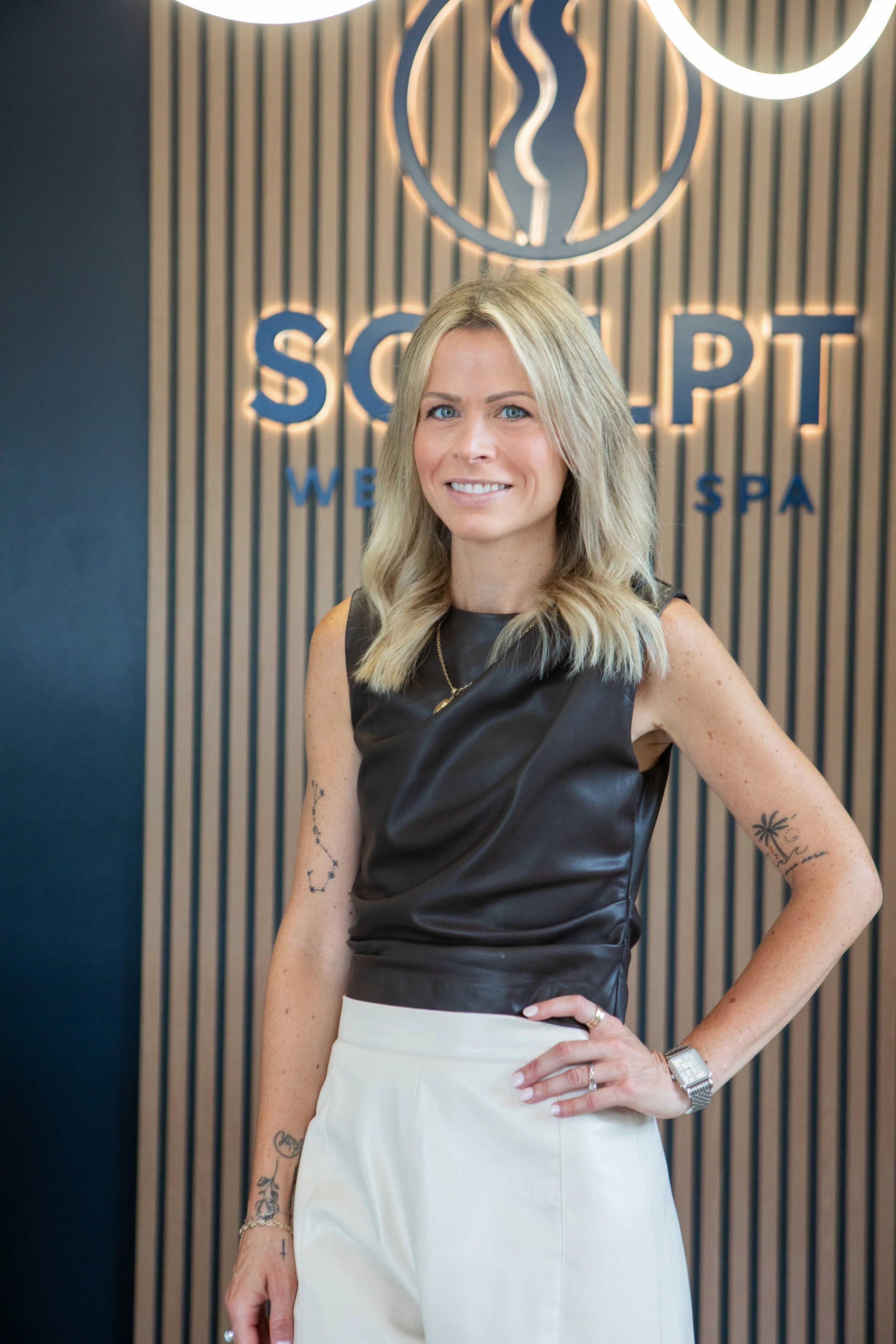 A woman with blonde hair, wearing a sleeveless black top and white pants, standing in front of a wall with a logo and text, smiling at the camera.