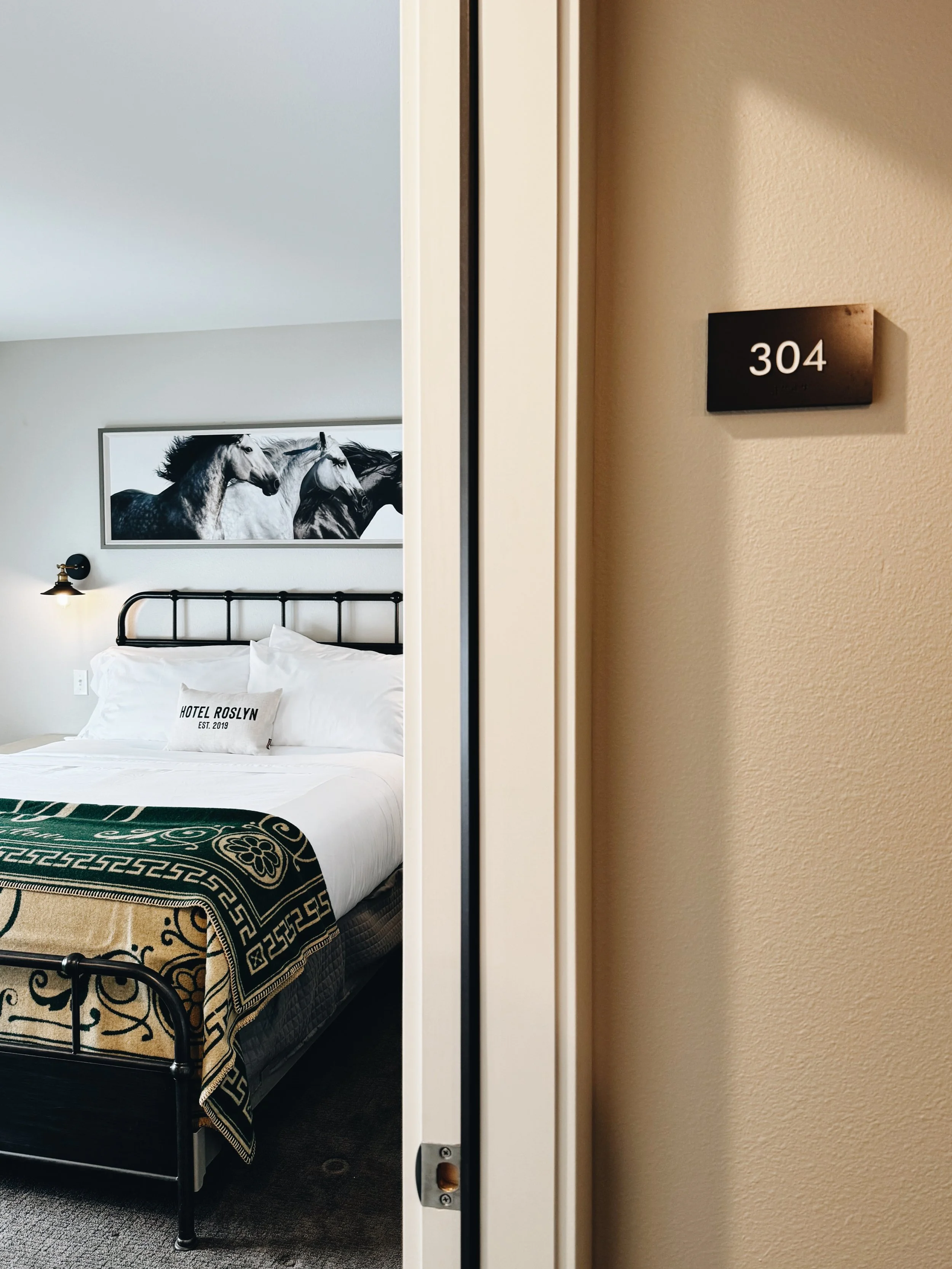 View of a hotel room with a black metal bed, a decorative blanket, and a pillow that reads 'HOTEL ROSLYN EST. 2019'; a black and white horse artwork on the wall, and a wall-mounted lamp.
