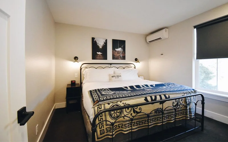 A 2nd Floor King Suite room at Hotel Roslyn, in Roslyn, WA. Hotel room with a metal bed frame, white bedding, and decorative pillows, two art prints above the bed, black bedside table, and black wall sconce lights.
