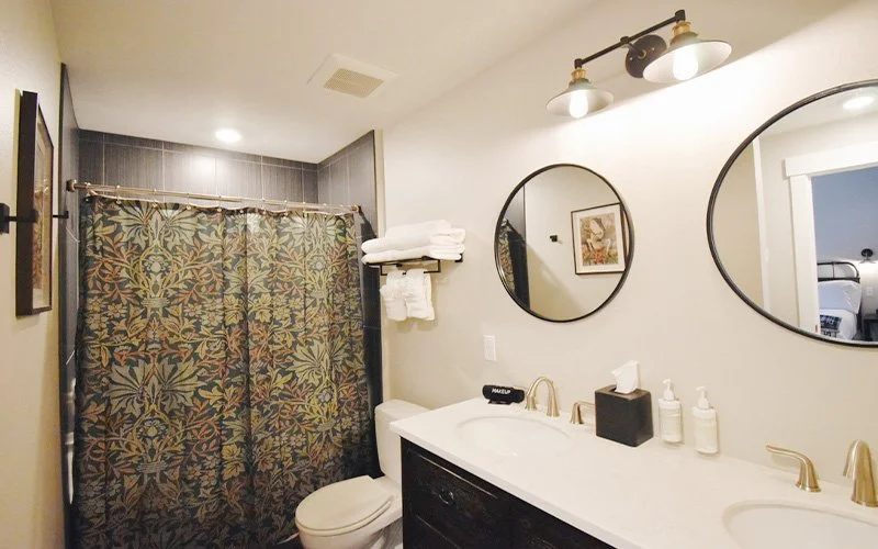 A King Patio room at Hotel Roslyn in Roslyn, WA. Bathroom with a shower area covered by a patterned curtain, a toilet, a white countertop with soap dispensers, tissue box, and two round mirrors, with wall-mounted lights above.