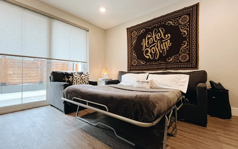 A King Patio room at Hotel Roslyn in Roslyn, WA. A hotel room with a large bed, a sofa, and a nightstand with a lamp. There is a wall with a large decorative sign that says 'Hotel Roslyn.' The room has a window with blinds and wooden flooring.