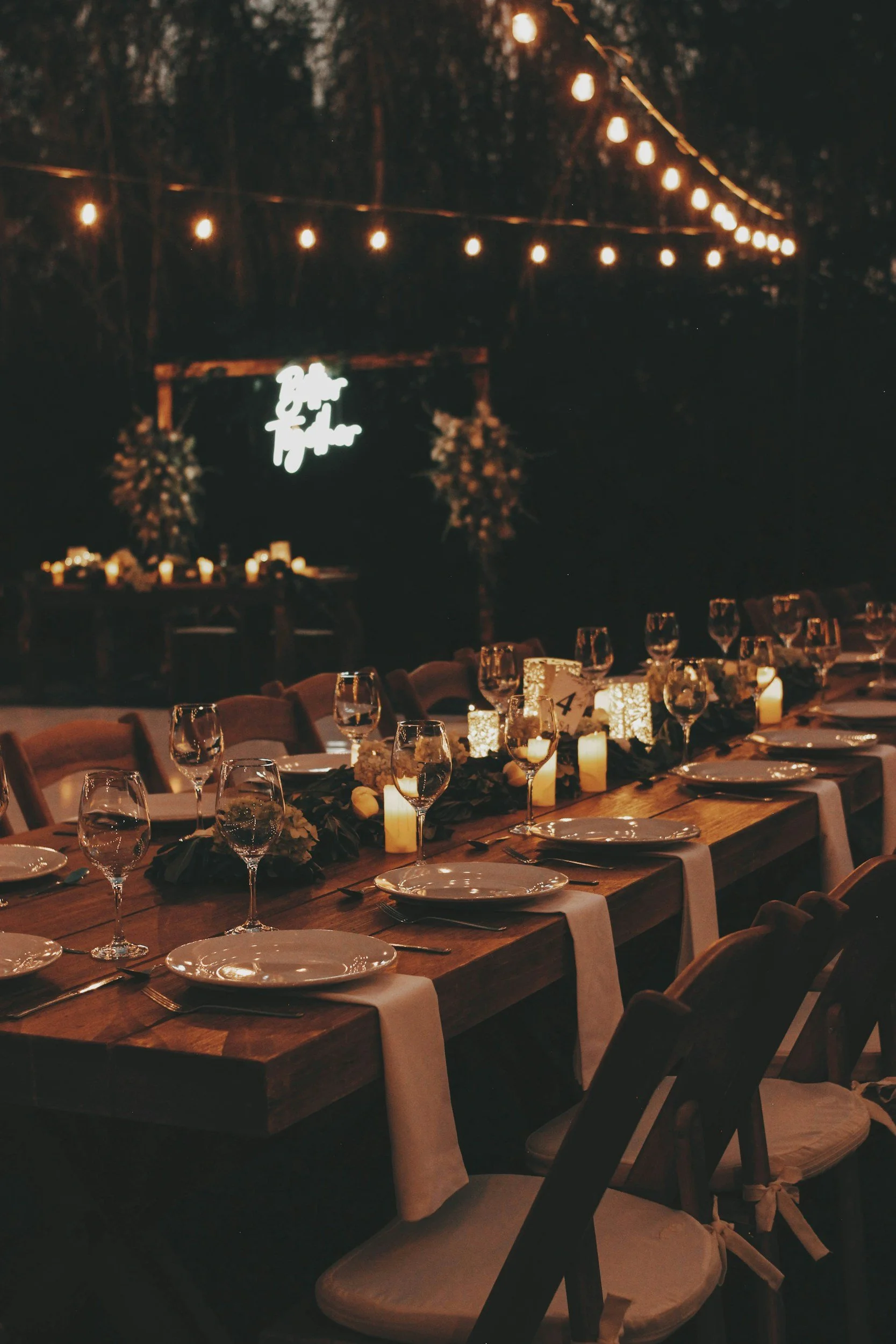 A long wooden dinner table set for an event with white napkins, wine glasses, and candles, decorated with flowers and greenery. String lights overhead and a neon sign in the background read 'Drink and T
