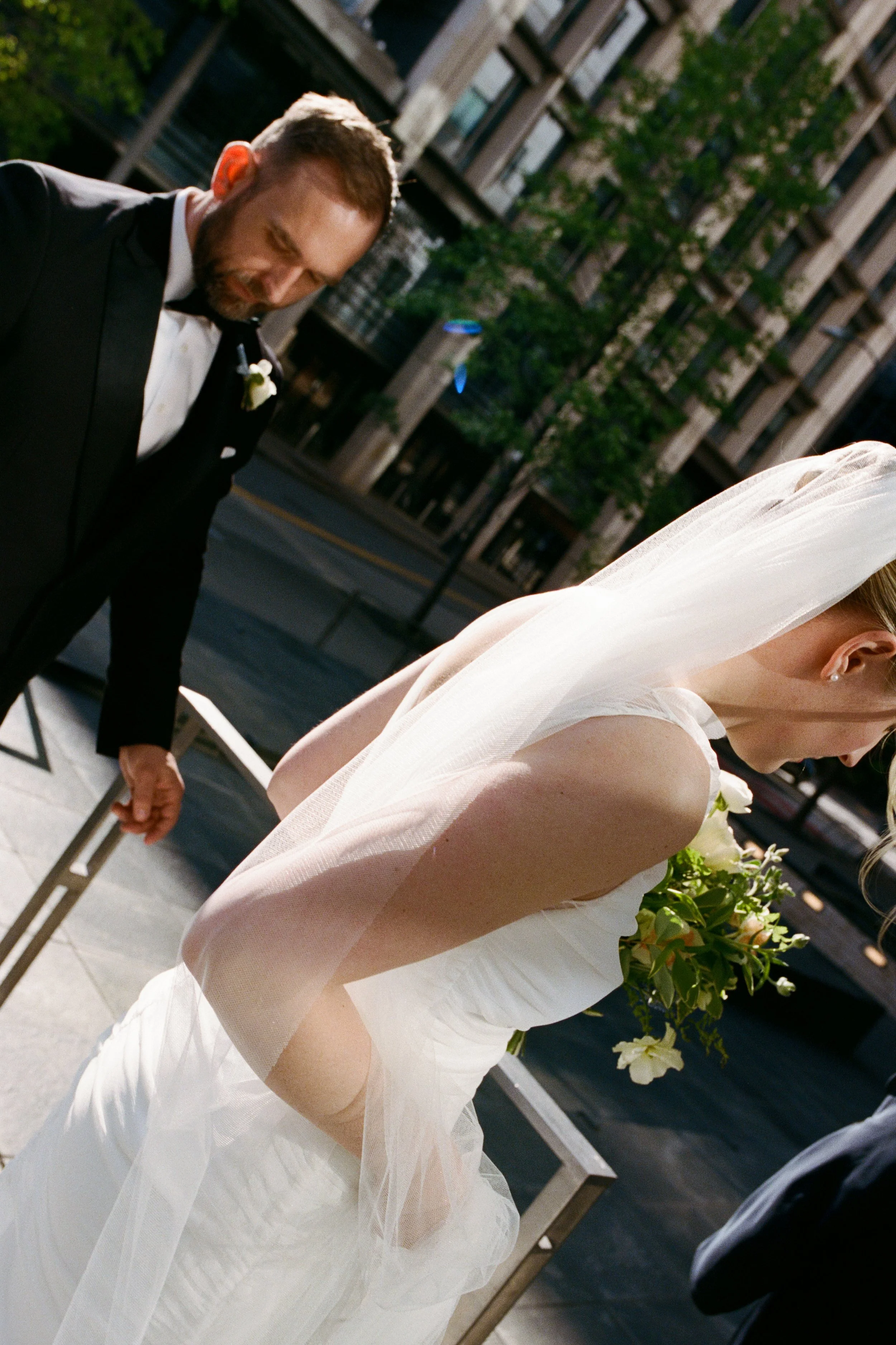 A bride in a white wedding dress holding a bouquet of flowers, and a groom in a black tuxedo with a boutonniere, standing outdoors in an urban setting during what appears to be a wedding ceremony.