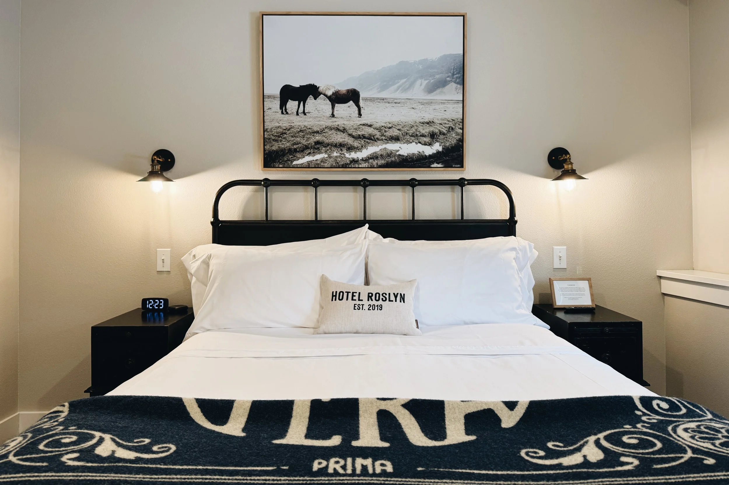 A hotel room with a bed, two black nightstands, and wall-mounted lamps. Above the bed is a framed picture of two horses on a beach. There is a pillow with the text "HOTEL ROSLYN EST. 2019" and a black blanket with white text at the foot of the bed. A small digital clock showing 12:23 is on the left nightstand.