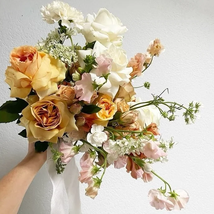 A hand holding a bouquet of various roses, lisianthus, and other flowers in shades of peach, white, and pink against a plain white wall.