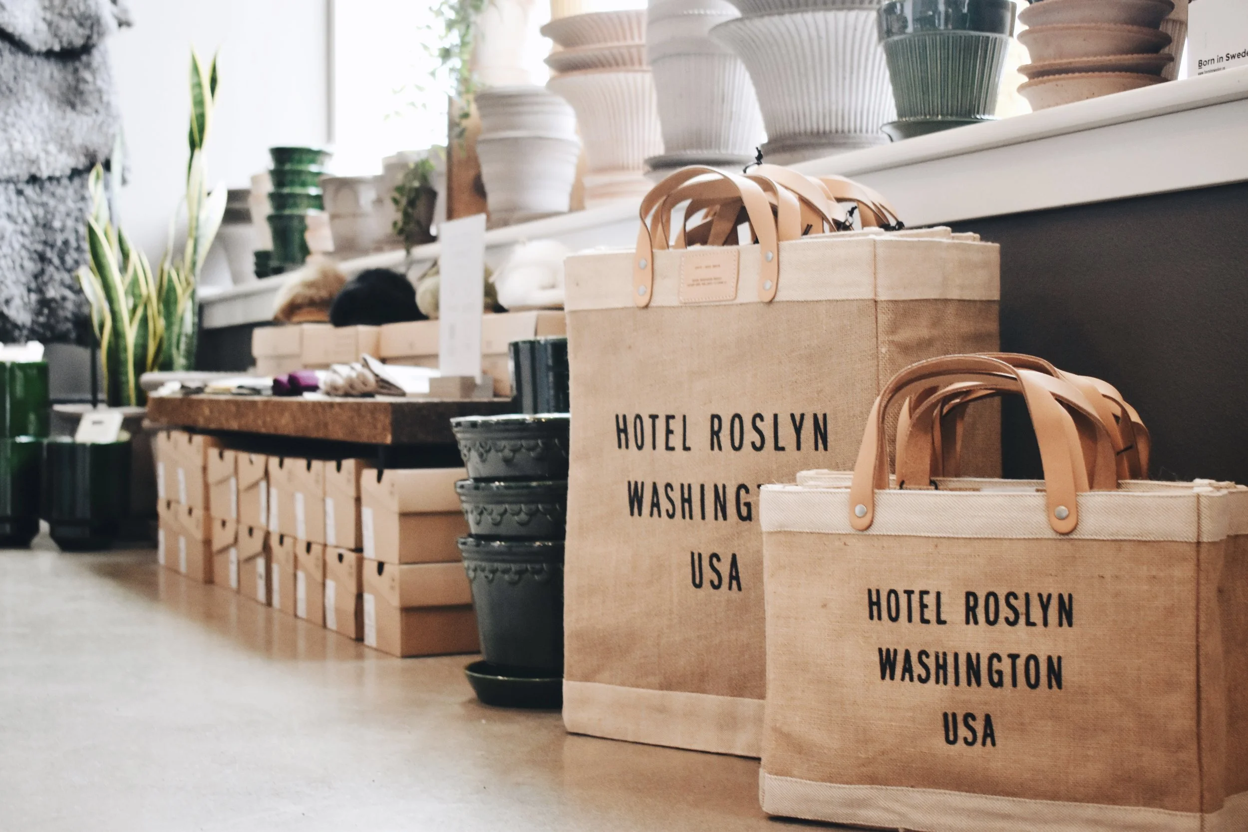 Rows of shopping bags made of tan fabric with leather handles, labeled 'Hotel Roslyn Washington USA,' placed on a wooden floor in a store with various decorative items and vases on shelves in the background.