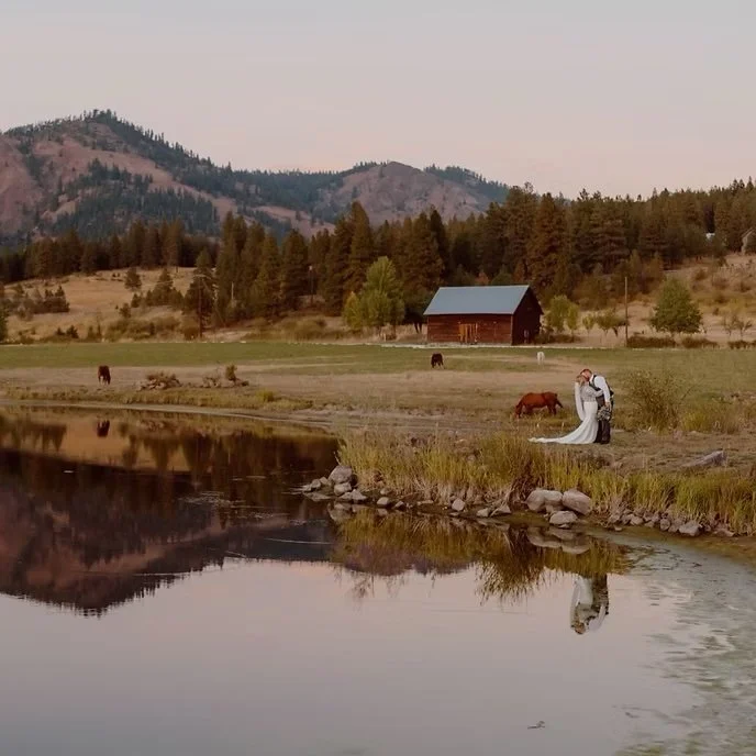 A couple in wedding attire standing by a river on a farm, with horses grazing, a barn, trees, and mountains in the background, and their reflection visible in the water.