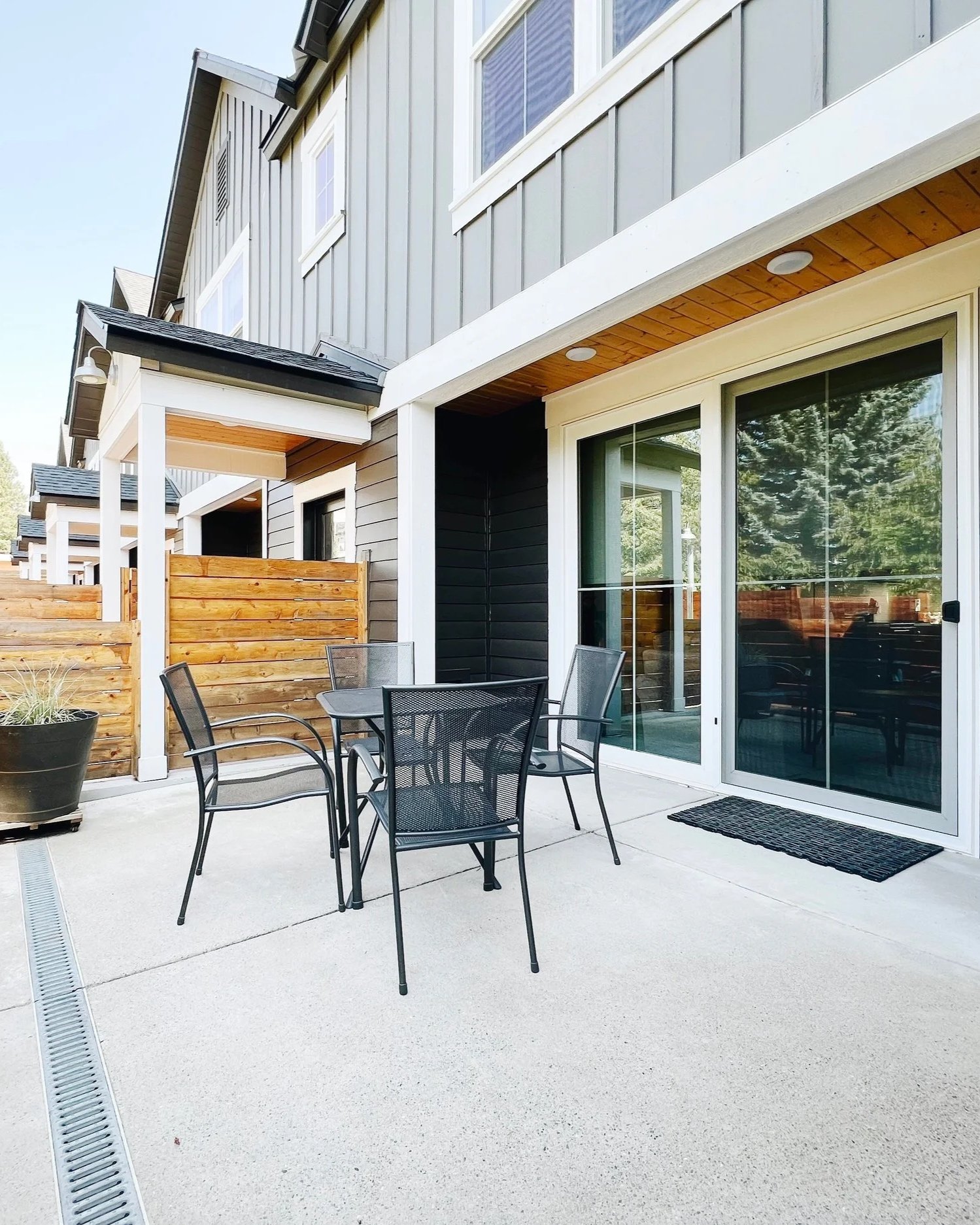 Apartment patio with black metal chairs around a table, large sliding glass door, potted plant, wooden privacy fence, and modern house exterior with gray siding and white trim.