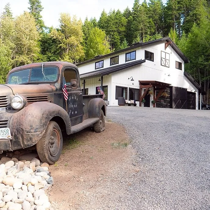 Old rusty black truck outside a modern white house with black accents, surrounded by green trees.