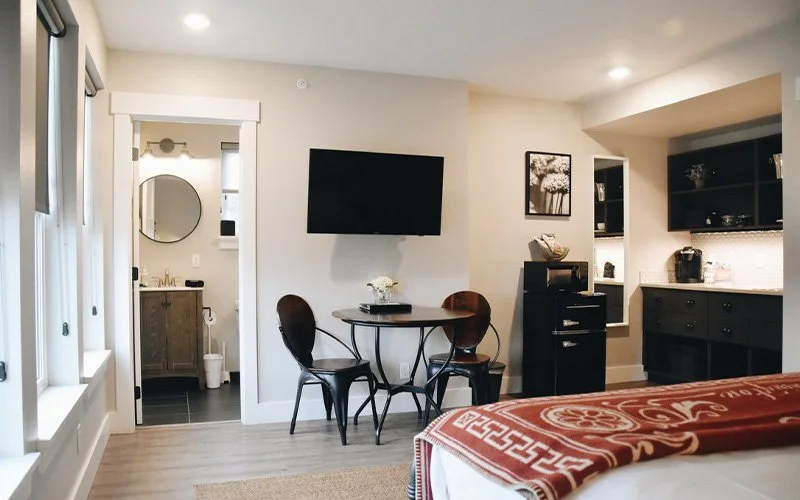 Living room with a wall-mounted TV, a small round dining table with two chairs, and a kitchen area in the background. A doorway leads to a bathroom with a mirror and sink.
