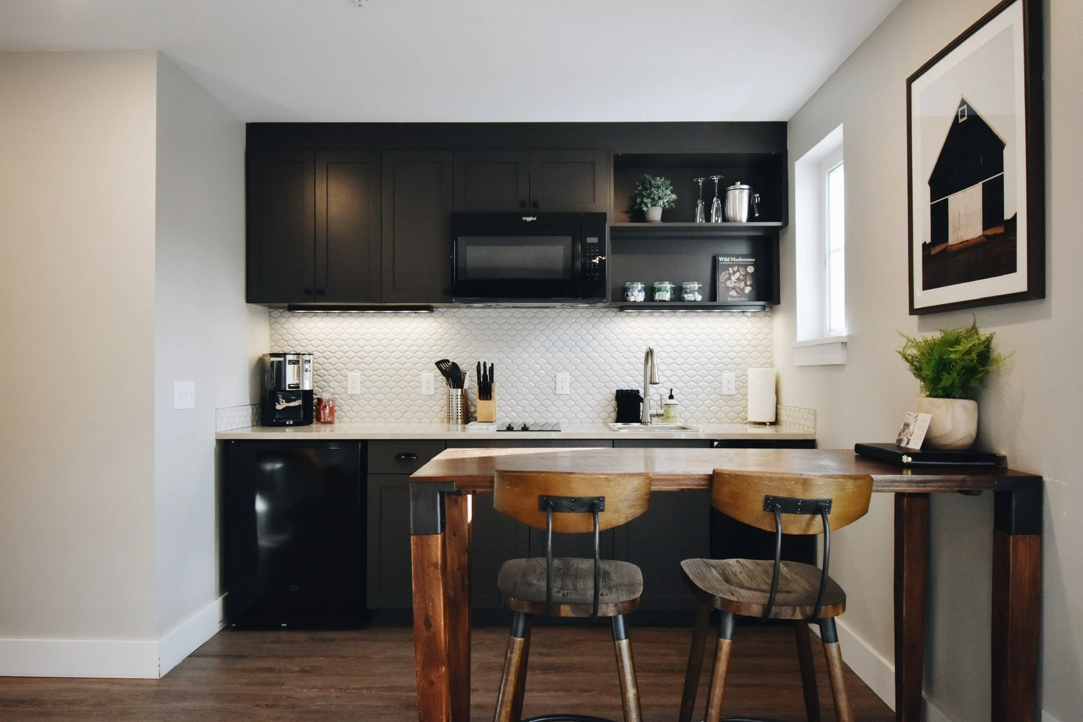 Modern kitchen with black cabinetry, white textured backsplash, wooden dining table, and chairs, decorated with potted plants and kitchen appliances, illuminated by natural light from a window.
