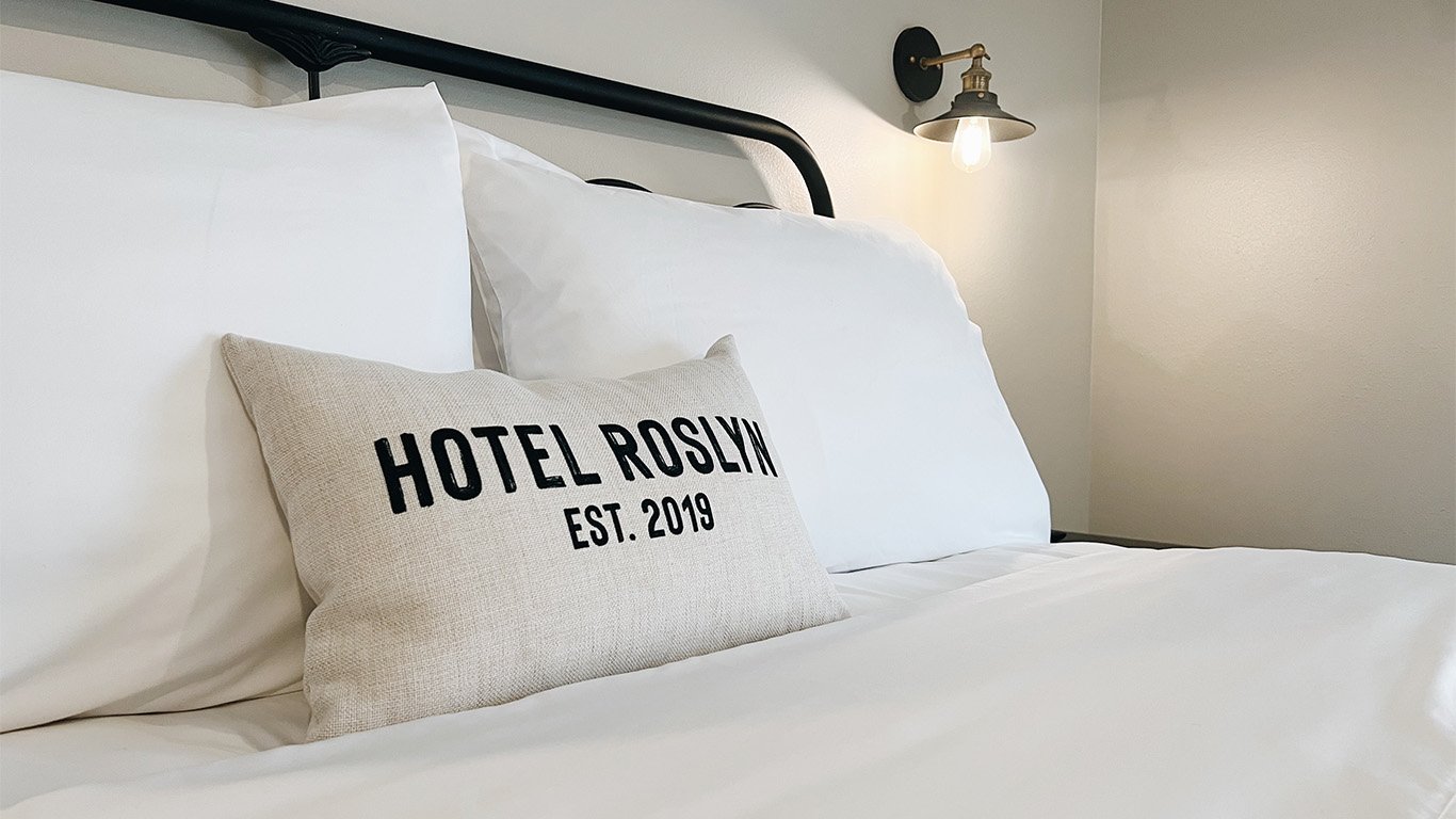 A neatly made bed with white sheets, a beige pillow with black text reading "HOTEL ROSLYN EST. 2019," and a black metal headboard. A wall-mounted light fixture is turned on in the background.