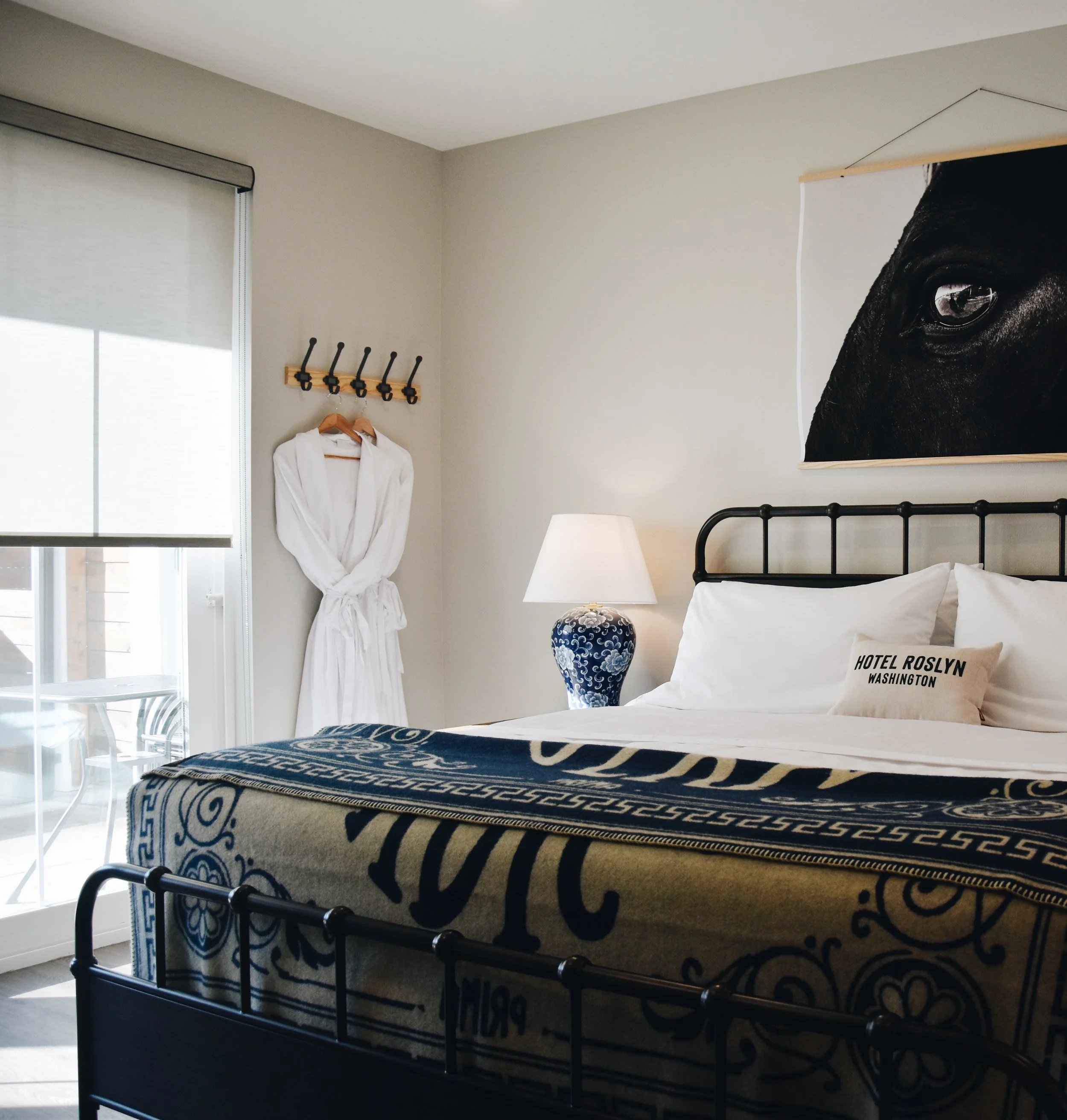 Hotel room with a bed, a bedside lamp, a black and white blanket, a framed black paw print on the wall, a white bathrobe hanging on the wall, and a large window with blinds.