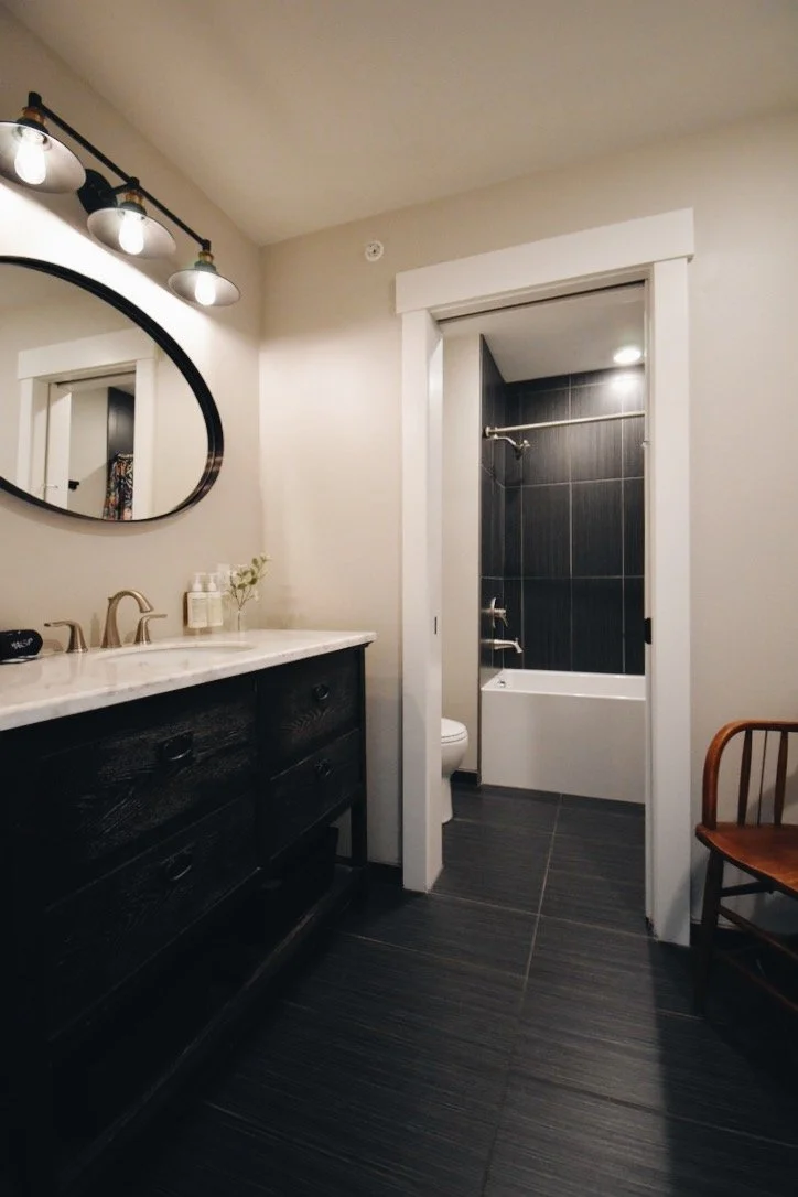 A 2nd Floor King Suite room at Hotel Roslyn, in Roslyn, WA. Modern bathroom with white walls, black vanity with marble countertop, oval mirror, black light fixture, and a separate shower and bathtub area with dark gray tiles.