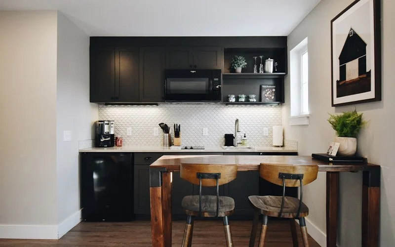 Kitchen area with dark upper cabinets, white backsplash, black appliances, and a wooden dining table with two chairs. There's a window on the right, artwork on the wall, and decorative items on the shelves.