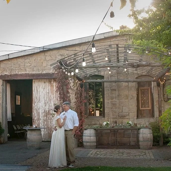 A couple is dancing closely in front of a rustic stone building with string lights overhead. The woman is in a white dress, and the man is in a light-colored shirt and tan pants. The scene is outdoors during the evening with warm lighting and greenery surroundings.